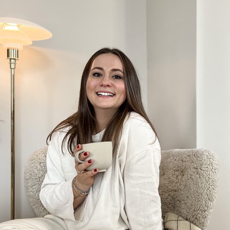 Lächelnde Frau im weißen Pullover hält eine beige Tasse, sitzt auf weichem Sessel in hellem Raum