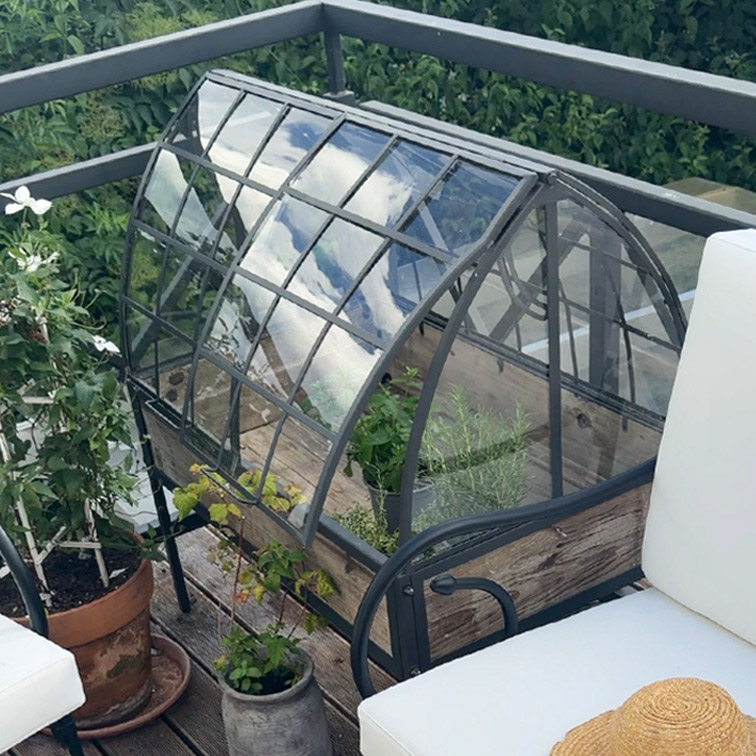 Kleine Gartengewächshaus mit Metallrahmen und Holzsockel auf Terrasse mit Pflanzen und Gartenmöbeln