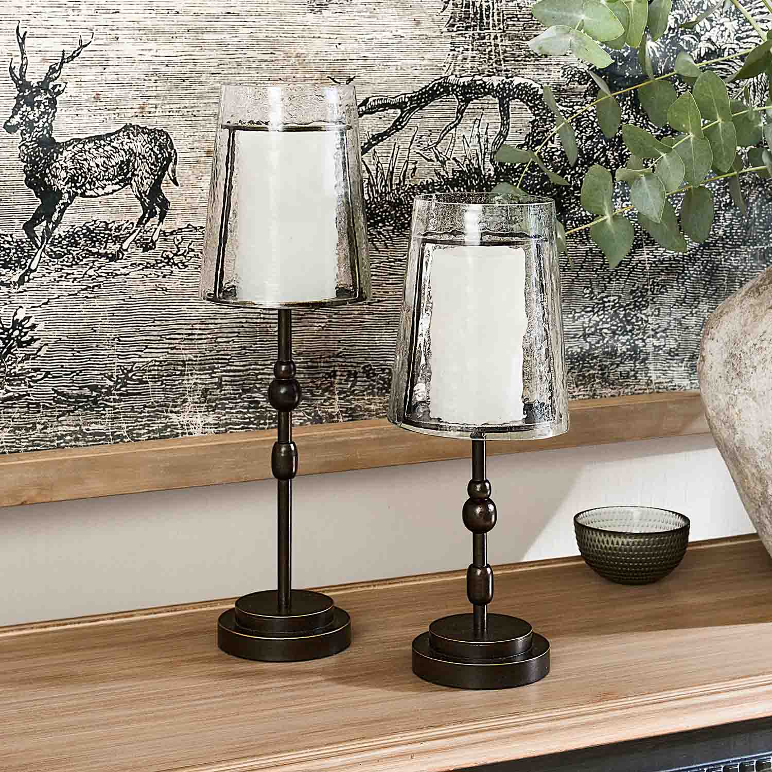 Zwei Metall-Kerzenhalter mit transparenten Glasschirmen und weißen Kerzen auf Holzablage