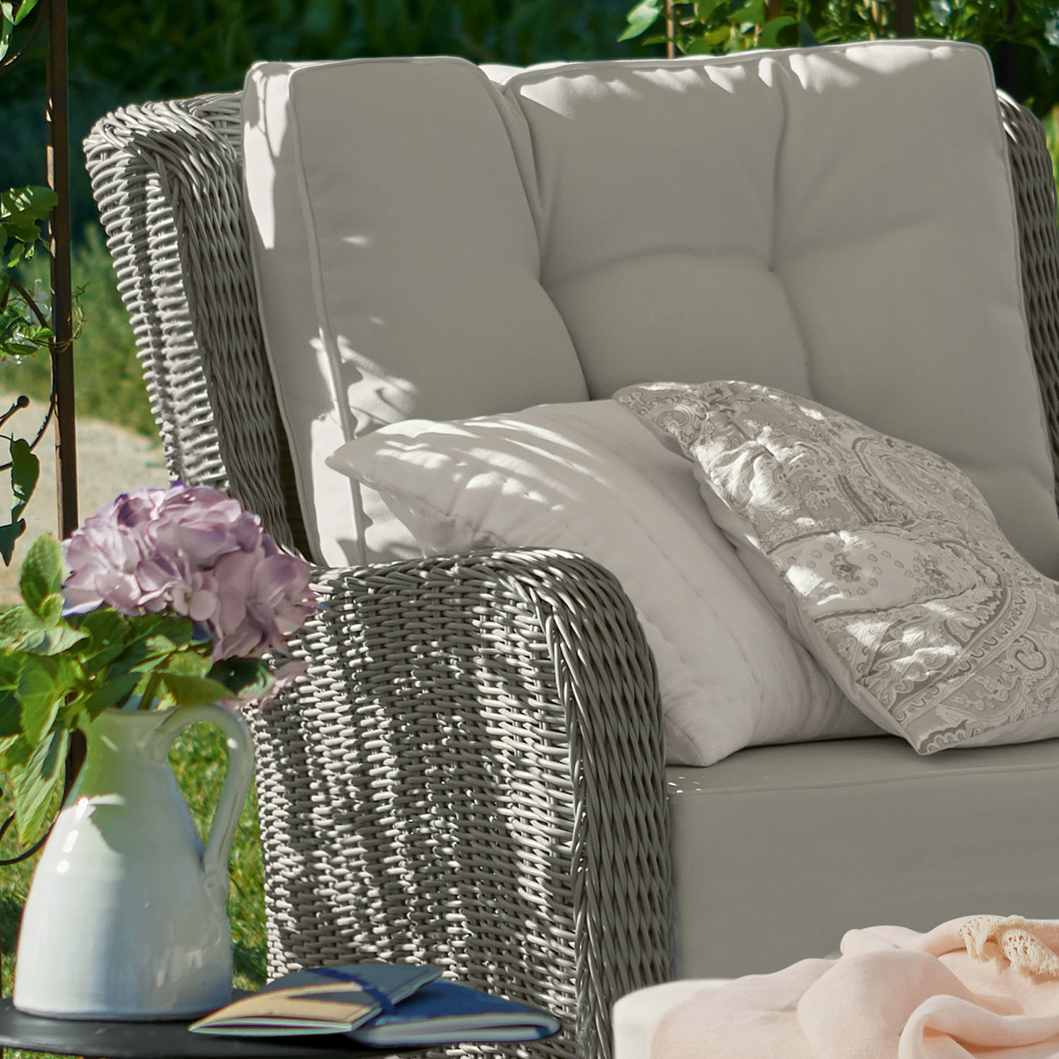 Graues Rattan-Sofa mit cremefarbenen Kissen und dekorativem Kissen im Garten mit Blumen