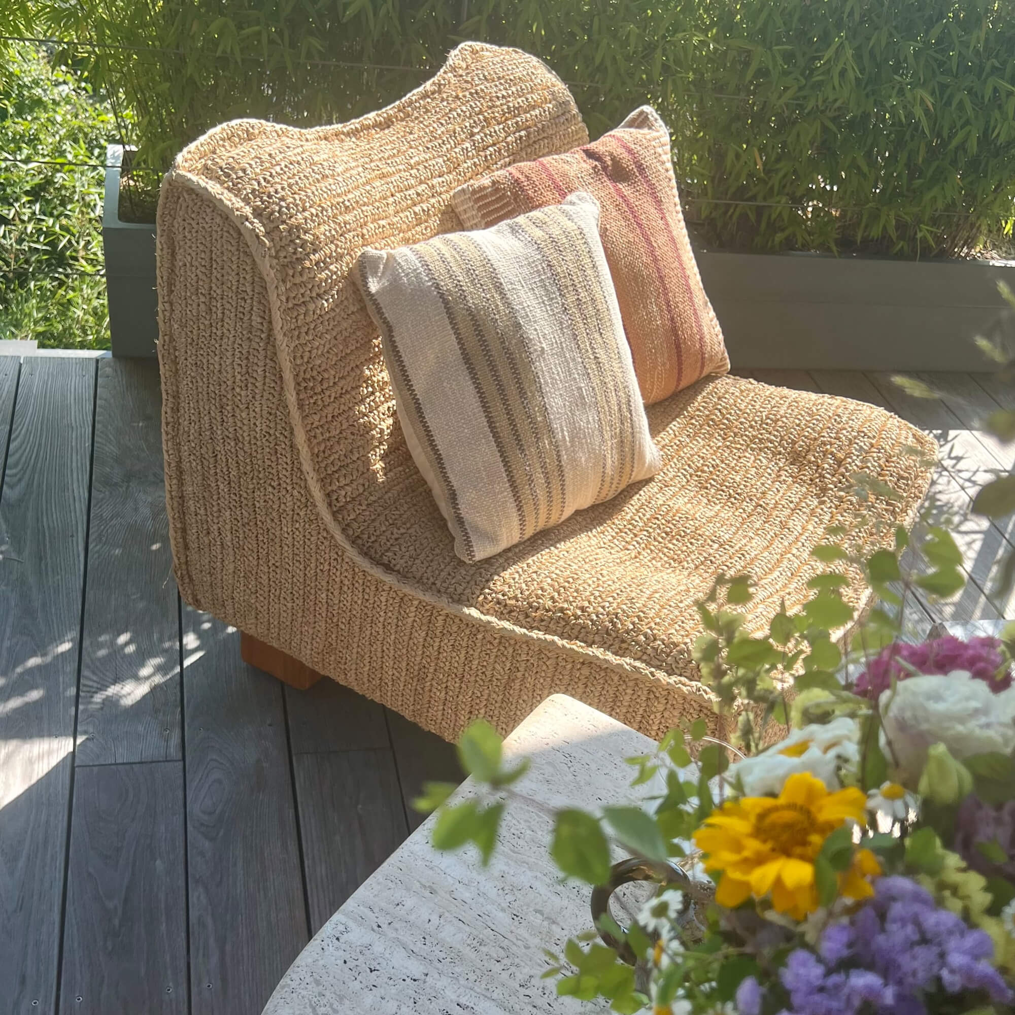 Beiger Sessel aus geflochtenem Material mit zwei dekorativen Kissen auf Terrasse mit Blumen