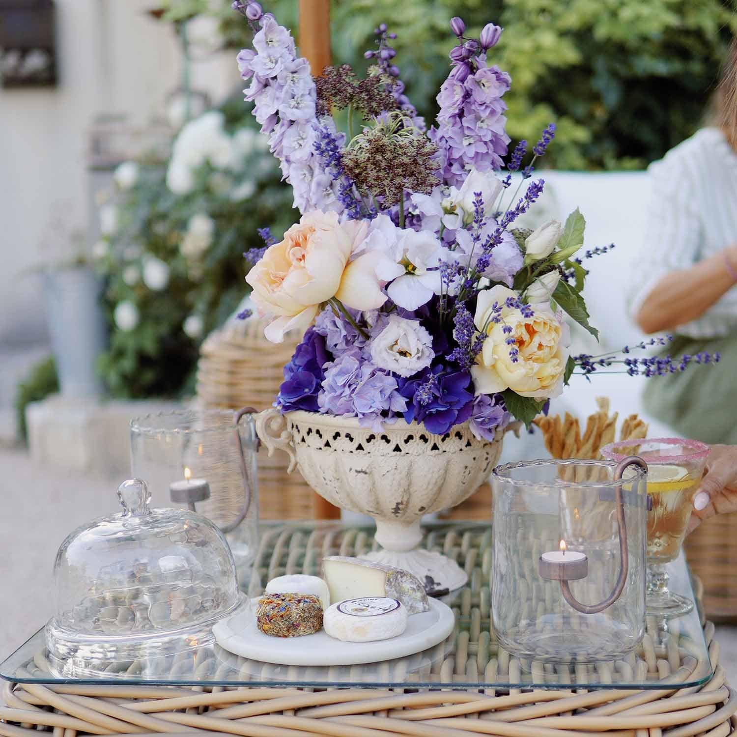 Strauß aus lila und cremefarbenen Blumen in weißer Keramikvase auf Glastisch mit Kerzen und Käseplatte