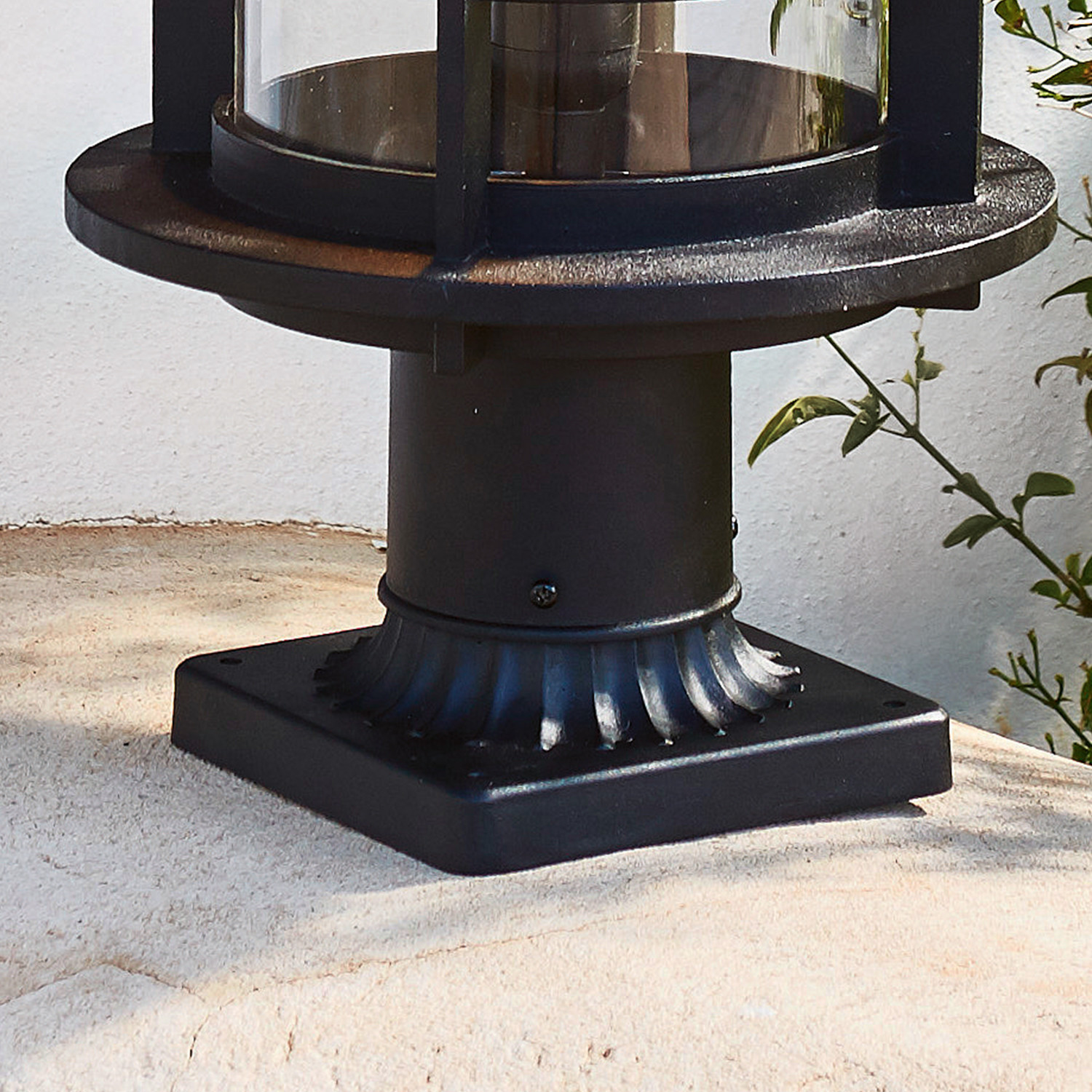 Schwarze Metallbasis einer Gartenlampe auf hellem Steinboden mit grünen Blättern im Hintergrund