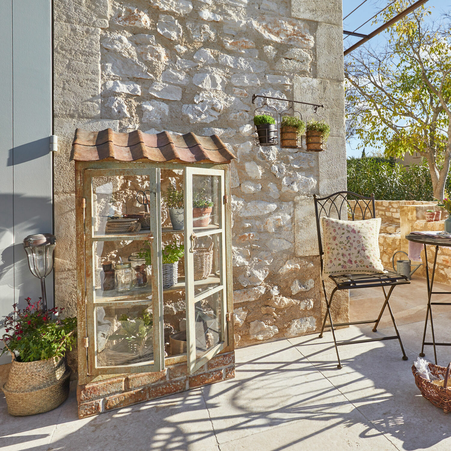 Metall-Gartenschrank mit Glastüren und Ziegeldach auf Terrasse vor Steinmauer im Sonnenlicht