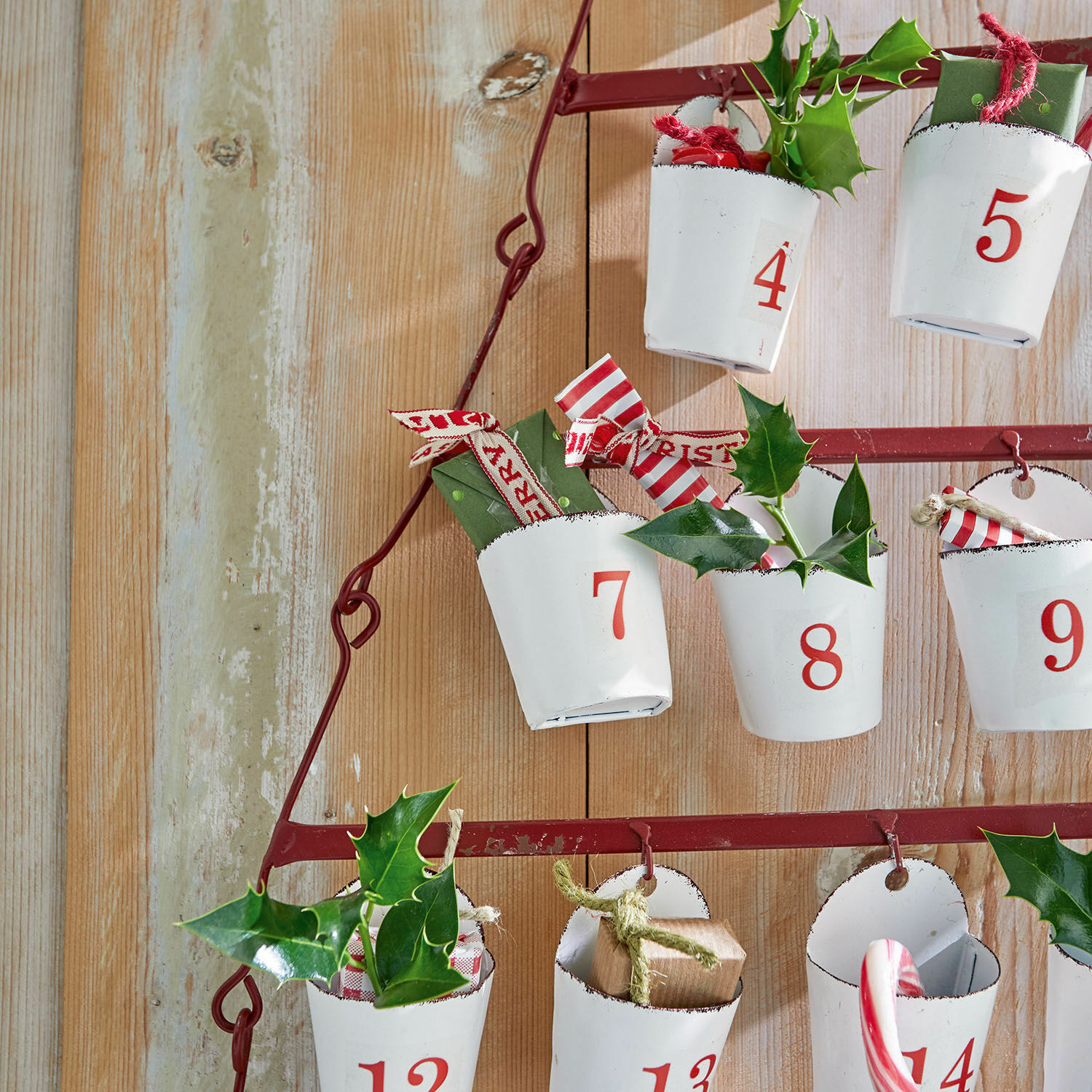 Adventskalender aus Metall mit weißen Bechern und roten Zahlen, dekoriert mit Stechpalmen und Geschenken