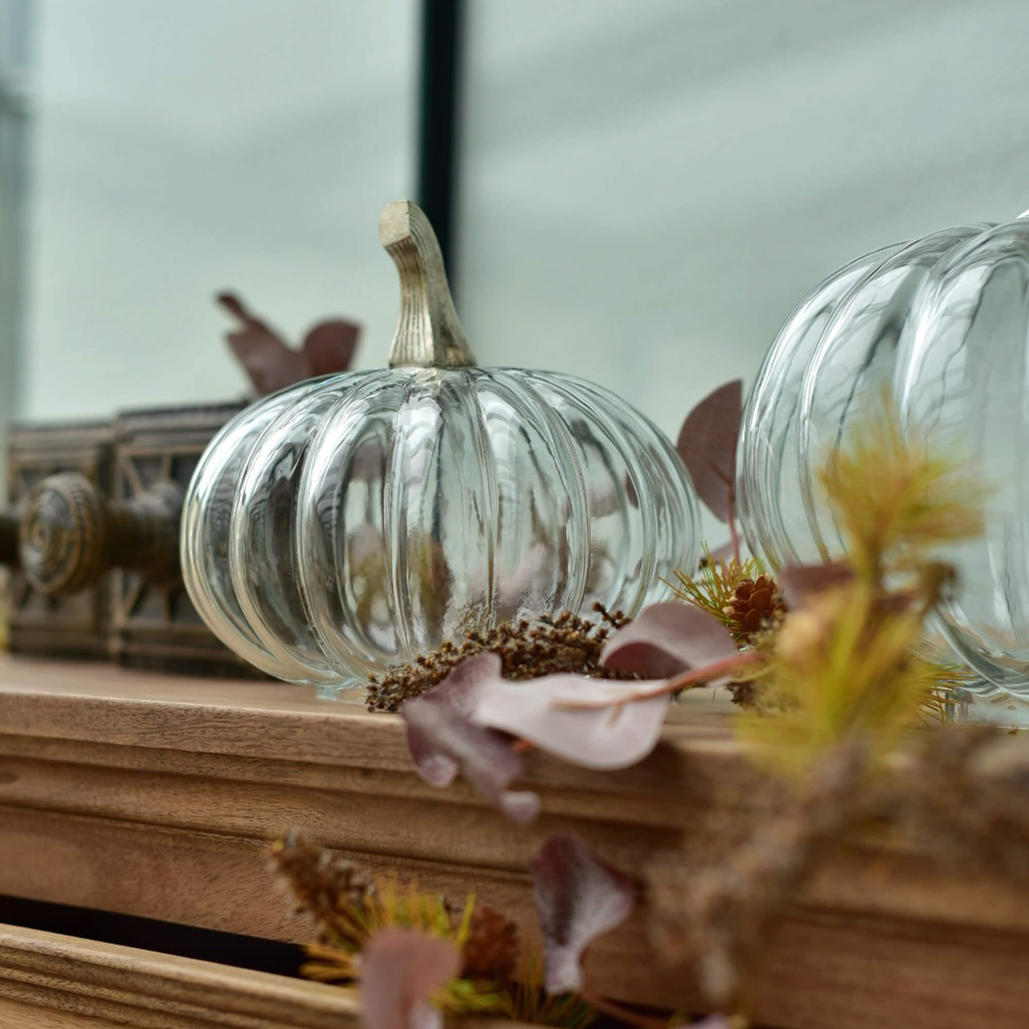 Transparanter Glas-Kürbis mit beigem Stiel auf Holzplatte mit herbstlicher Dekoration
