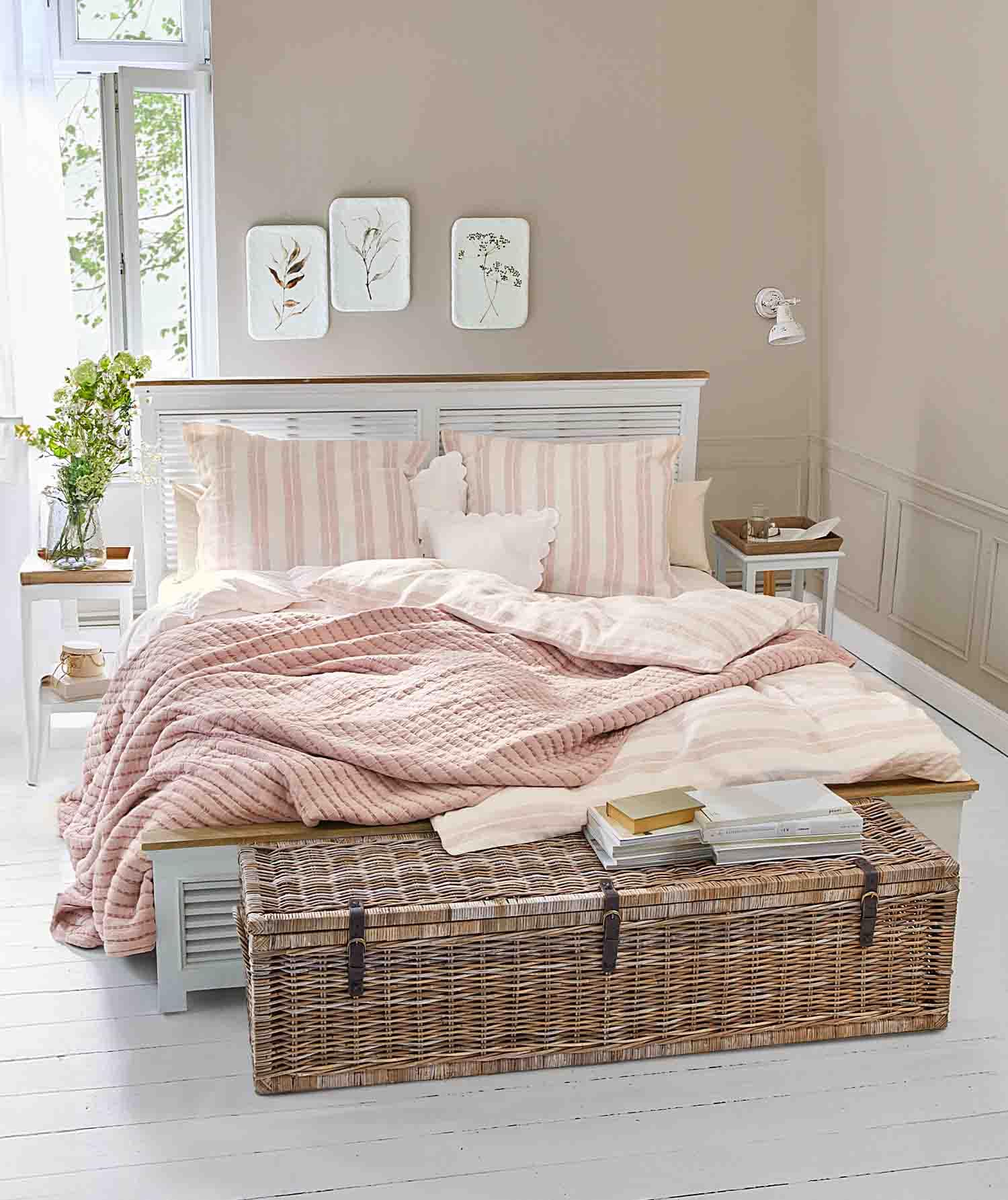 Gemütliches Schlafzimmer mit weißem Bett, rosa gesteppter Decke und einem Korbtruhe mit Büchern darauf