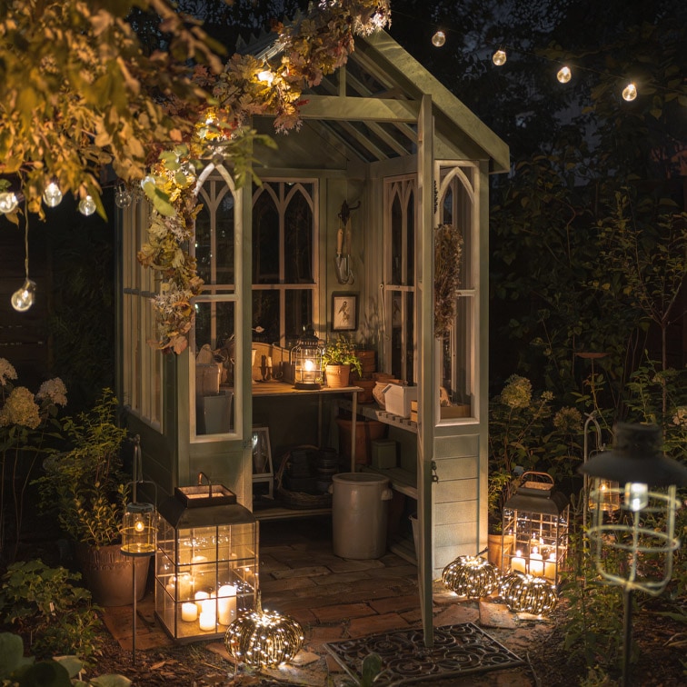 Beleuchtetes Holz-Gewächshaus mit dekorativen Laternen und Kerzen im Garten bei Nacht