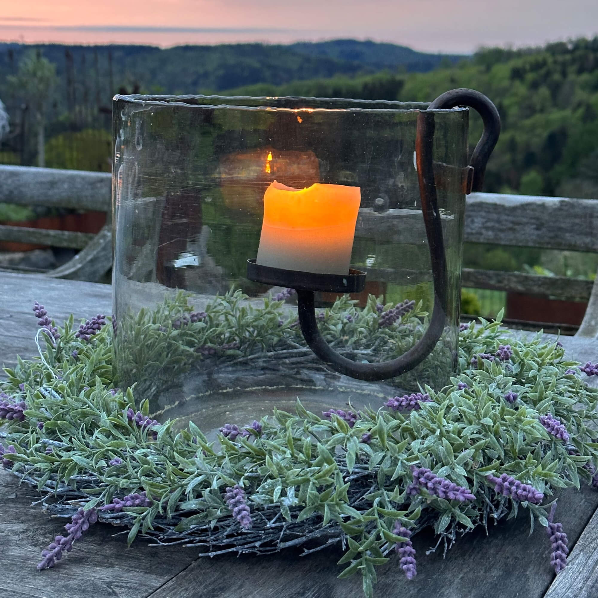 Glaswindlicht mit Metallhalter und grünem Kranz mit violetten Blüten auf Holztisch