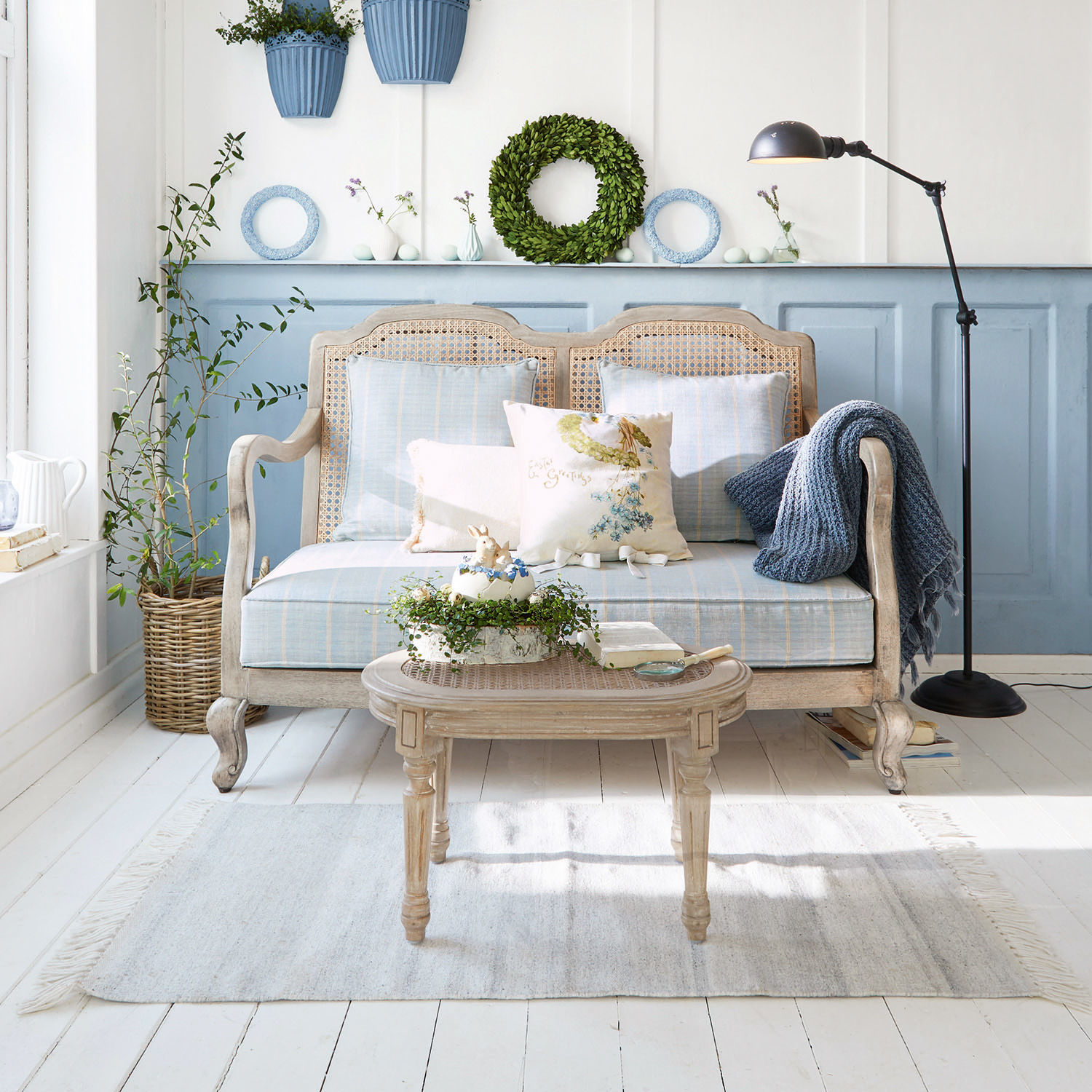 Sofa Ainsley Holz-Rattan-Sofa mit blauer Polsterung und Kissen in hellem Wohnzimmer mit Pflanzen und Stehlampe
