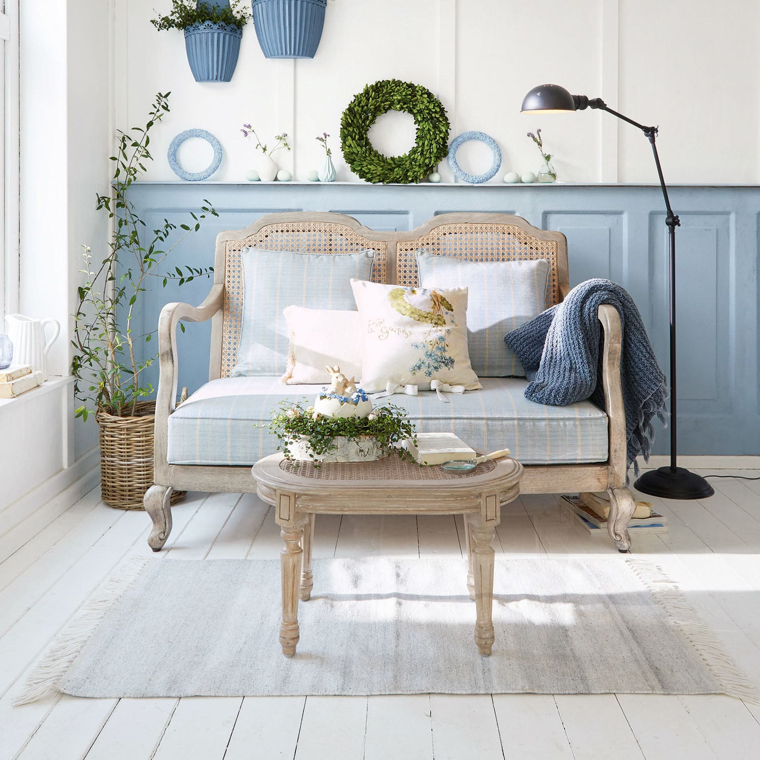 Sofa Ainsley Holz-Rattan-Sofa mit blauer Polsterung und Kissen in hellem Wohnzimmer mit Pflanzen und Stehlampe