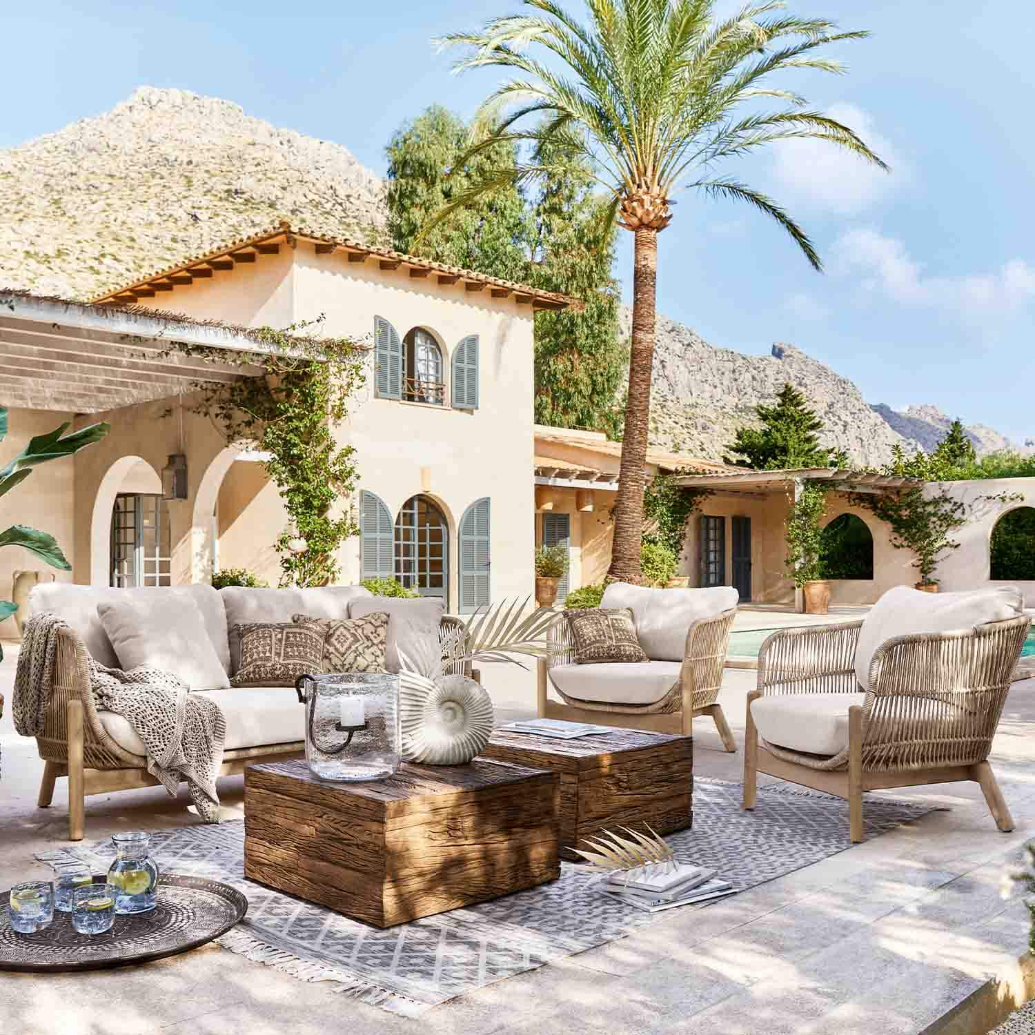 Outdoor-Möbelset aus hellem Rattan mit beigen Polstern auf Terrasse mit Palme und Bergblick