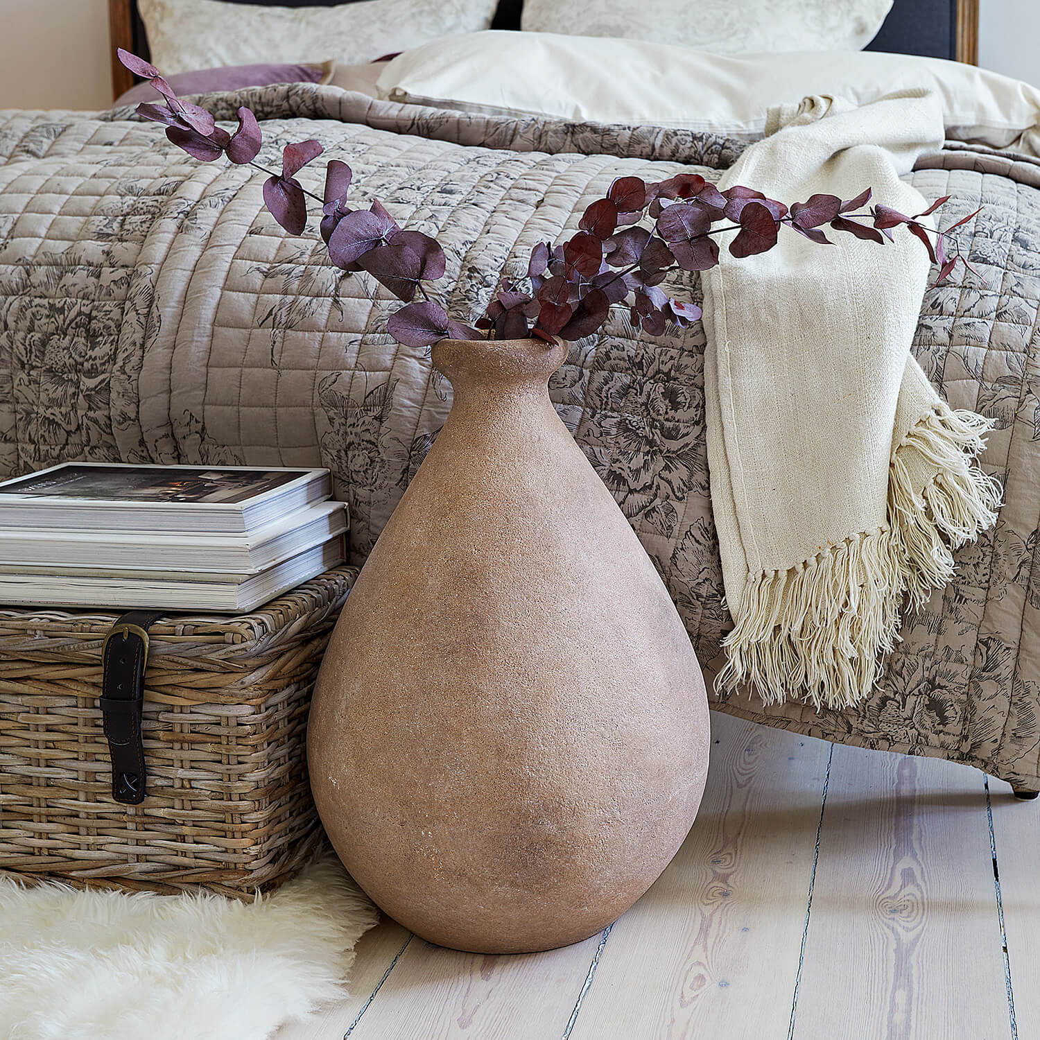 Große beige Keramikvase mit getrockneten Zweigen vor Bett und Weidenkorb