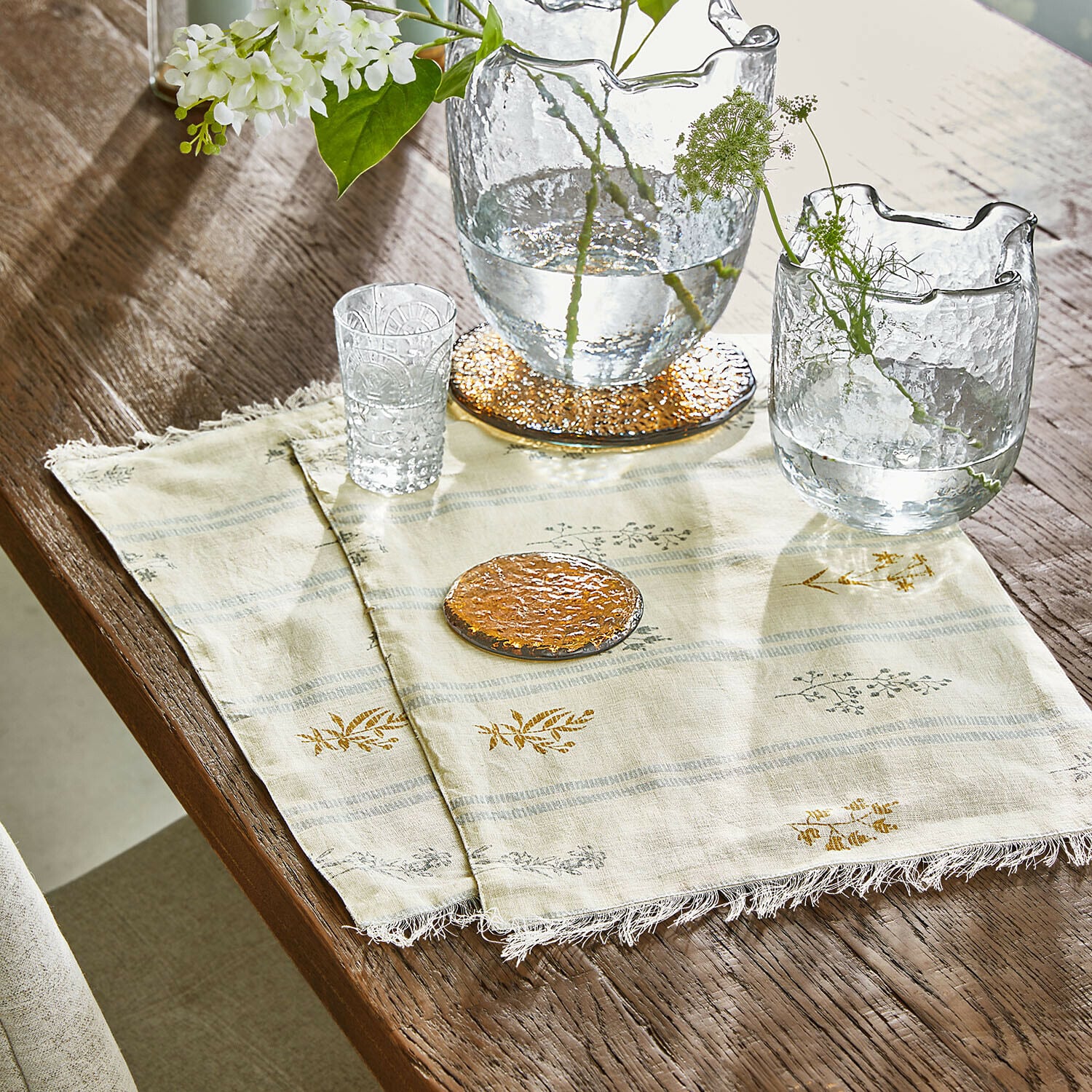 Transparente Glasvasen und Glas auf cremefarbenem Leinentuch mit Blumenmuster auf Holztisch