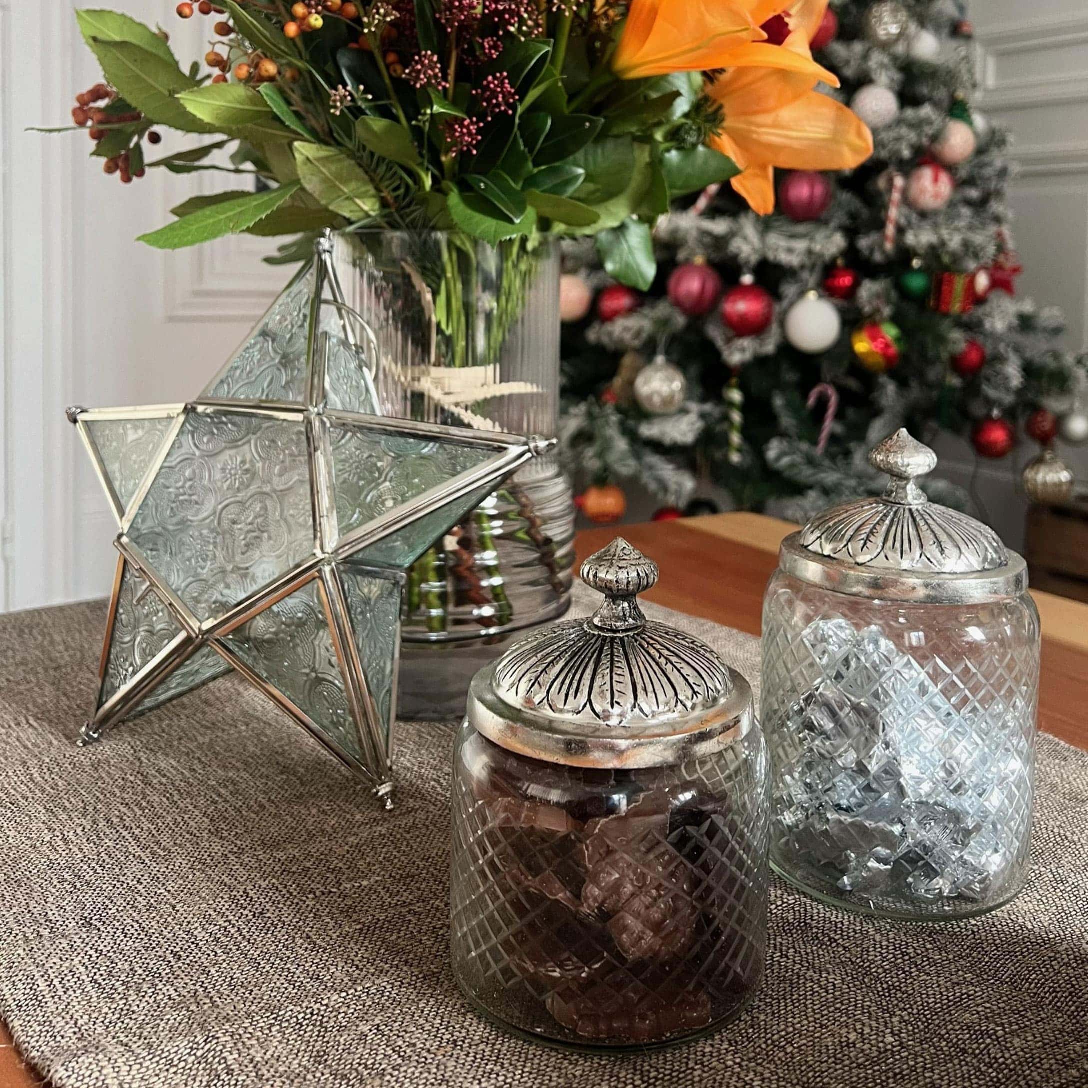 Gläser mit Metalldeckeln und dekorativer Stern auf Tisch mit Blumen und Weihnachtsbaum im Hintergrund