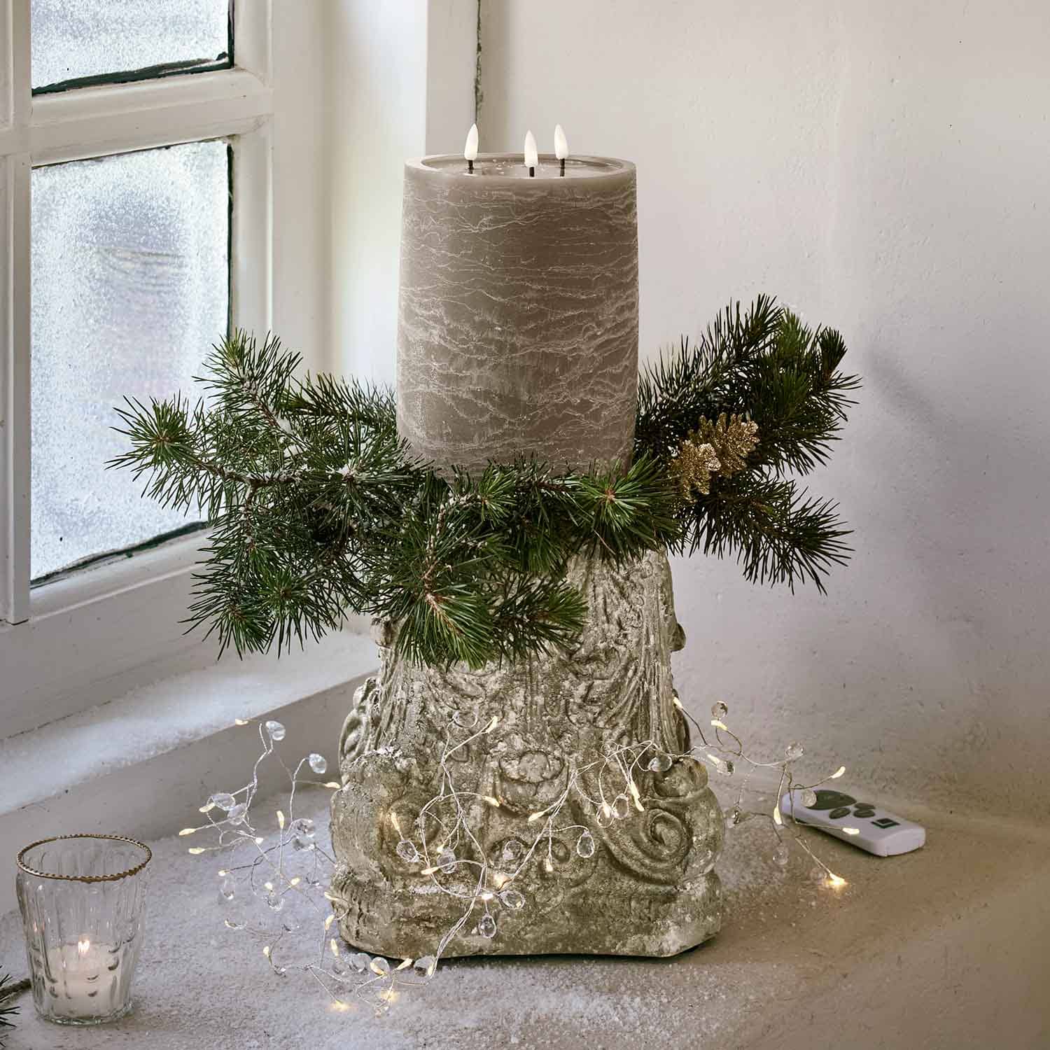 Graue Dreifachdochtkerze auf dekorativem Sockel mit grünen Zweigen und Weihnachtsdeko