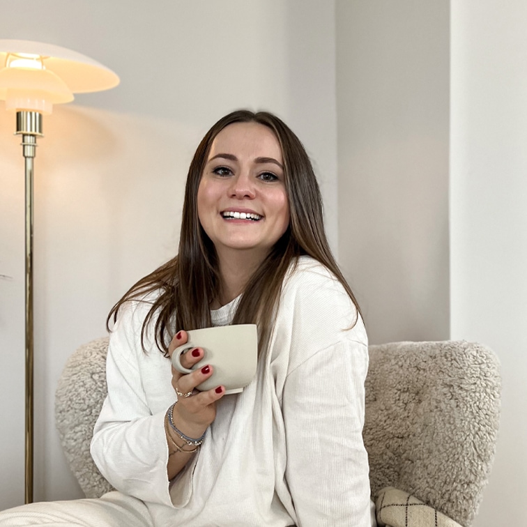 Lächelnde Frau im weißen Pullover hält eine beige Tasse, sitzt auf weichem Sessel in hellem Raum