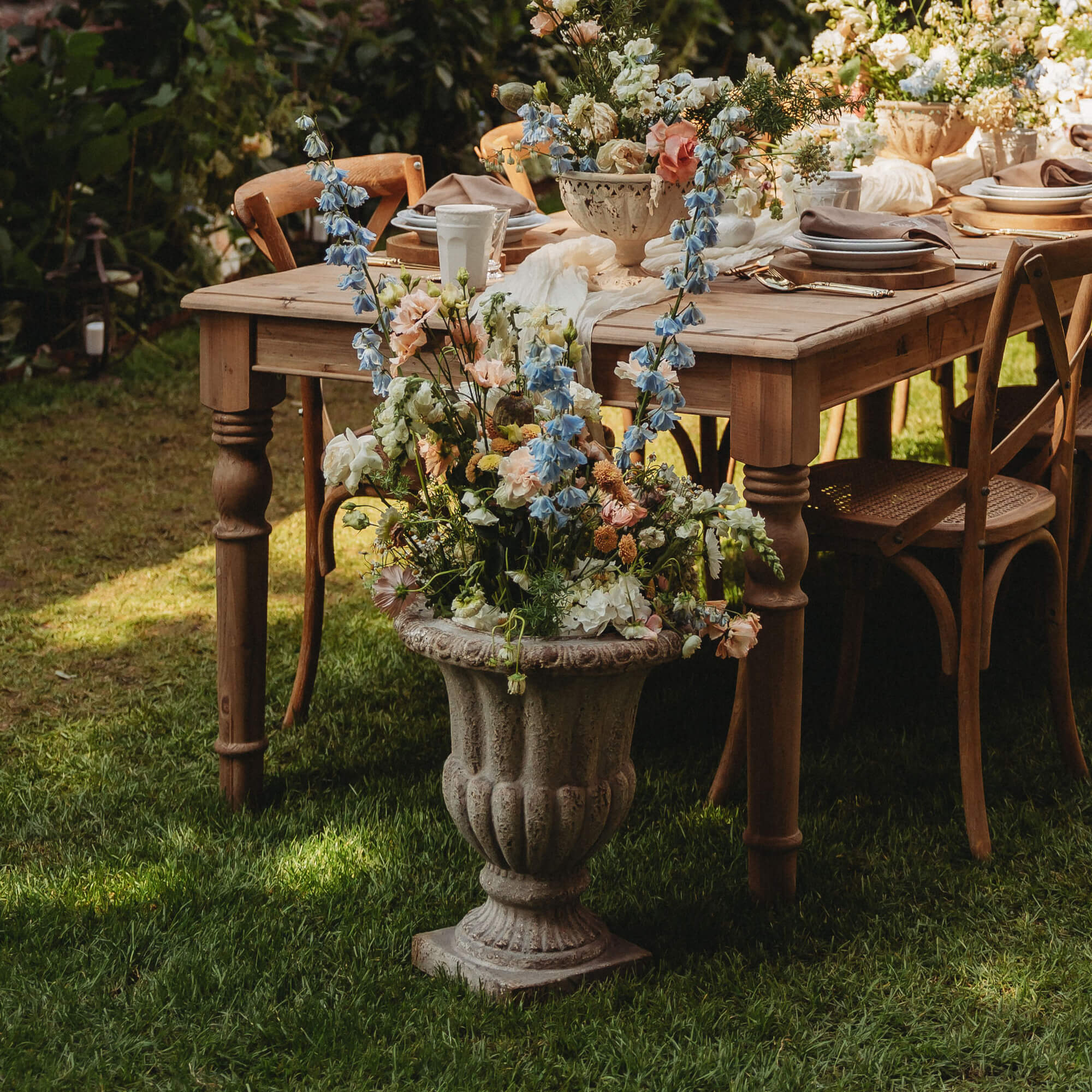 Große steinerne Blumenvase mit pastellfarbenen Blumen vor einem Holztisch und Stühlen im Garten