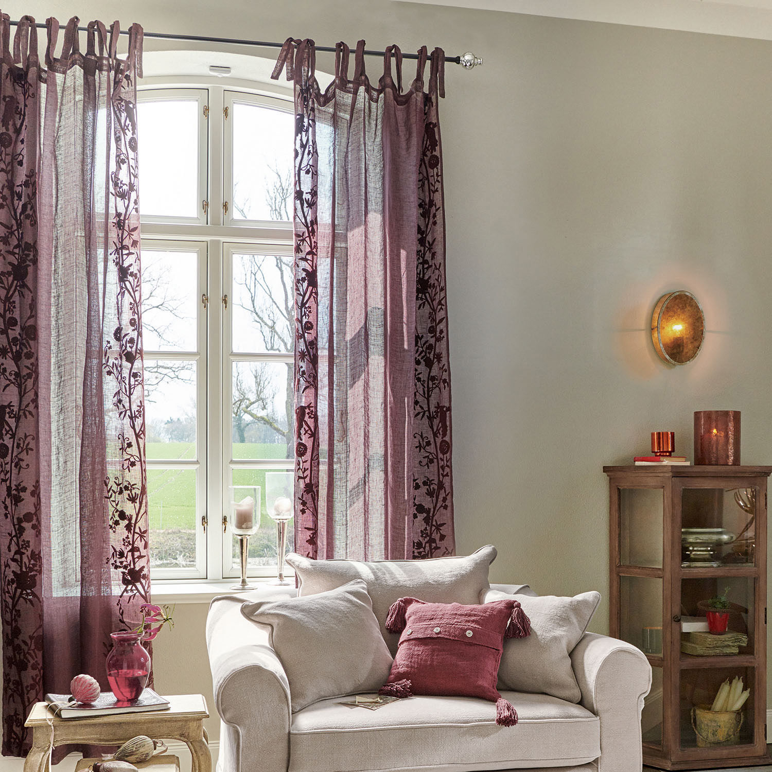 Gemütliches Wohnzimmer mit beigem Sofa und bordeauxroten, bestickten Vorhängen, Wohnaccessoires