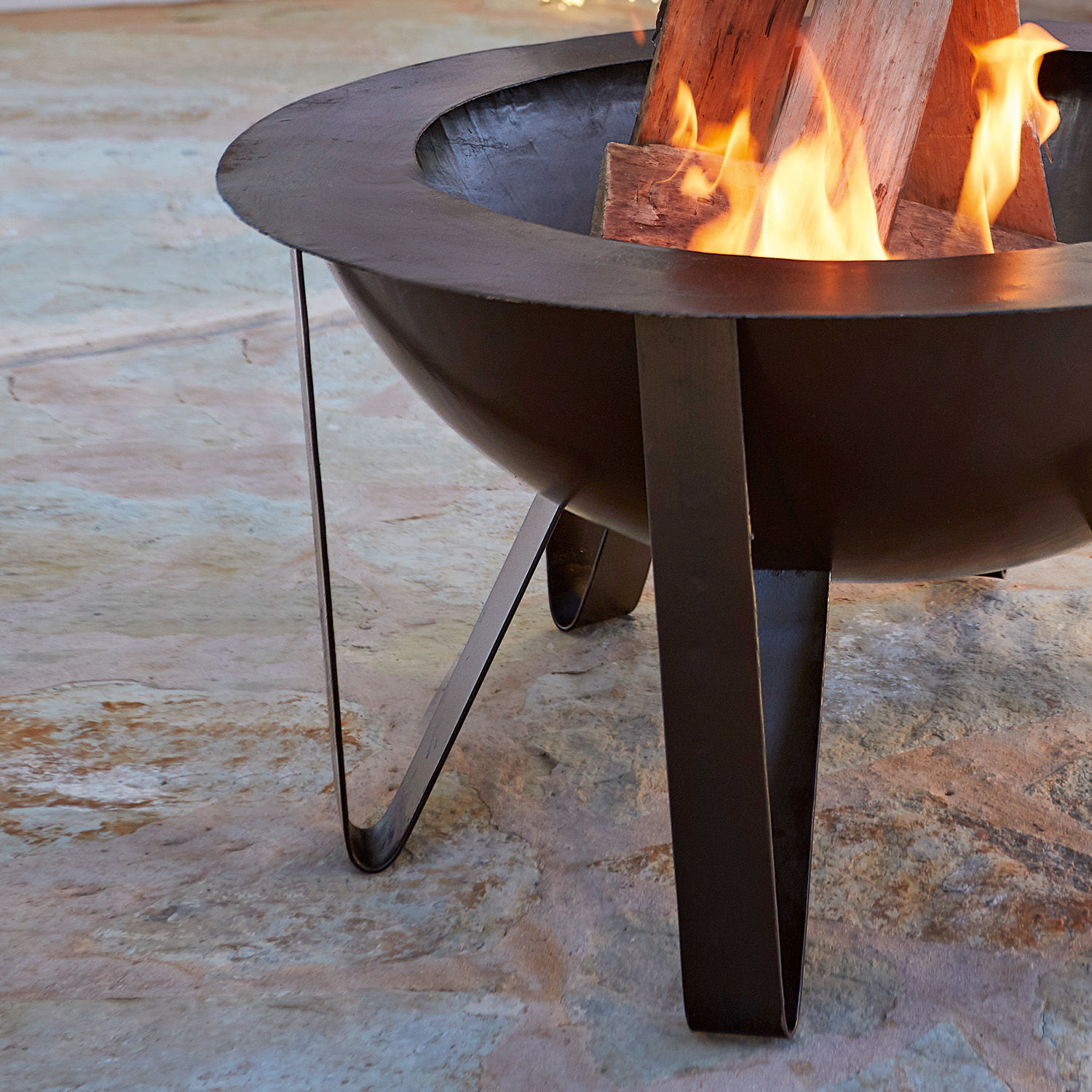 Schwarzes Metall-Feuerkorb mit brennendem Holz auf Steinboden im Garten