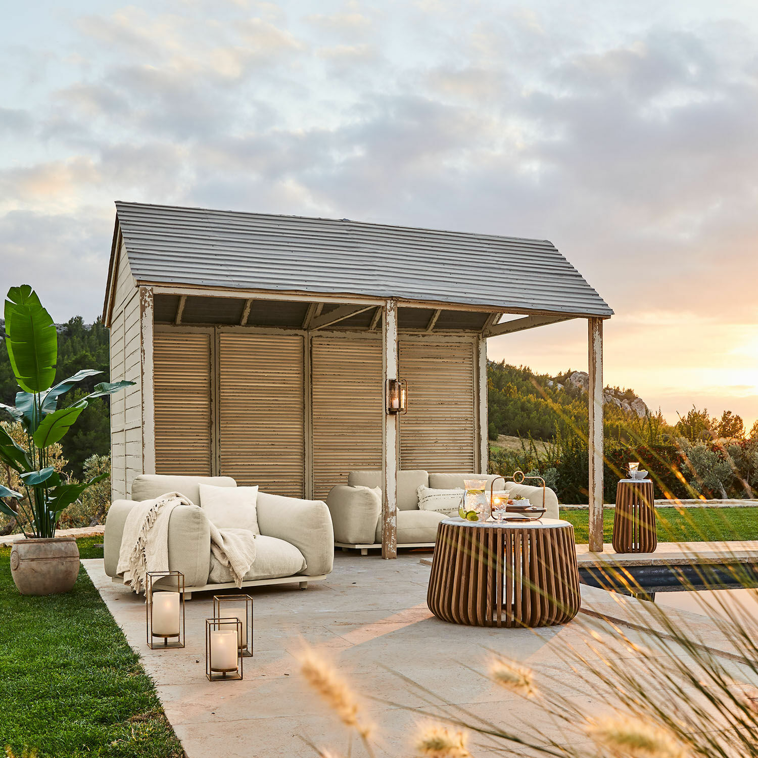 Gemütliche Gartenmöbel mit beigen Polstern und Holz-Tisch auf Terrasse neben Pool bei Sonnenuntergang