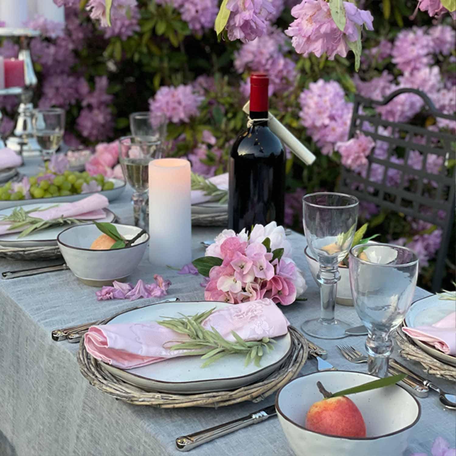 Eleganter gedeckter Tisch mit weißem Geschirr, rosa Servietten und rotem Wein in blühendem Garten