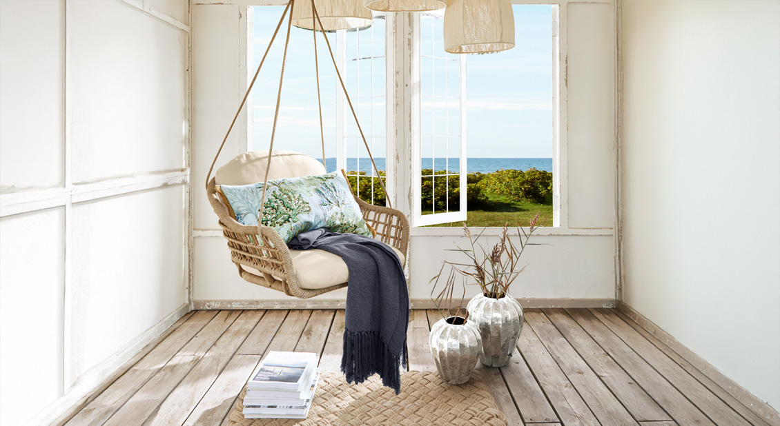 Hängesessel aus Korbgeflecht mit hellem Polster und maritimem Dekokissen in hellem Raum mit Meerblick