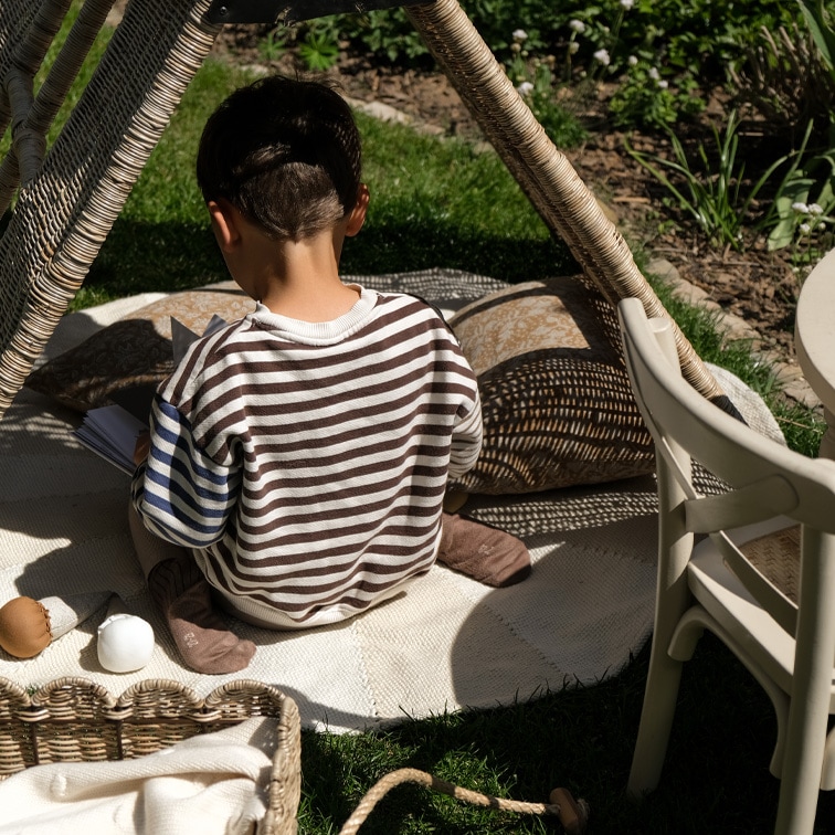 Kind im gestreiften Pullover sitzt auf Teppich unter Weidenzelt im Garten, entspannte Spielsituation