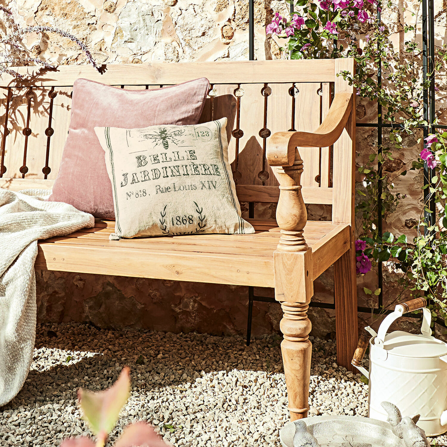 Helle Holz-Gartenbank mit pastellfarbenen Kissen auf Kiesboden, umgeben von Blumen und Gießkanne