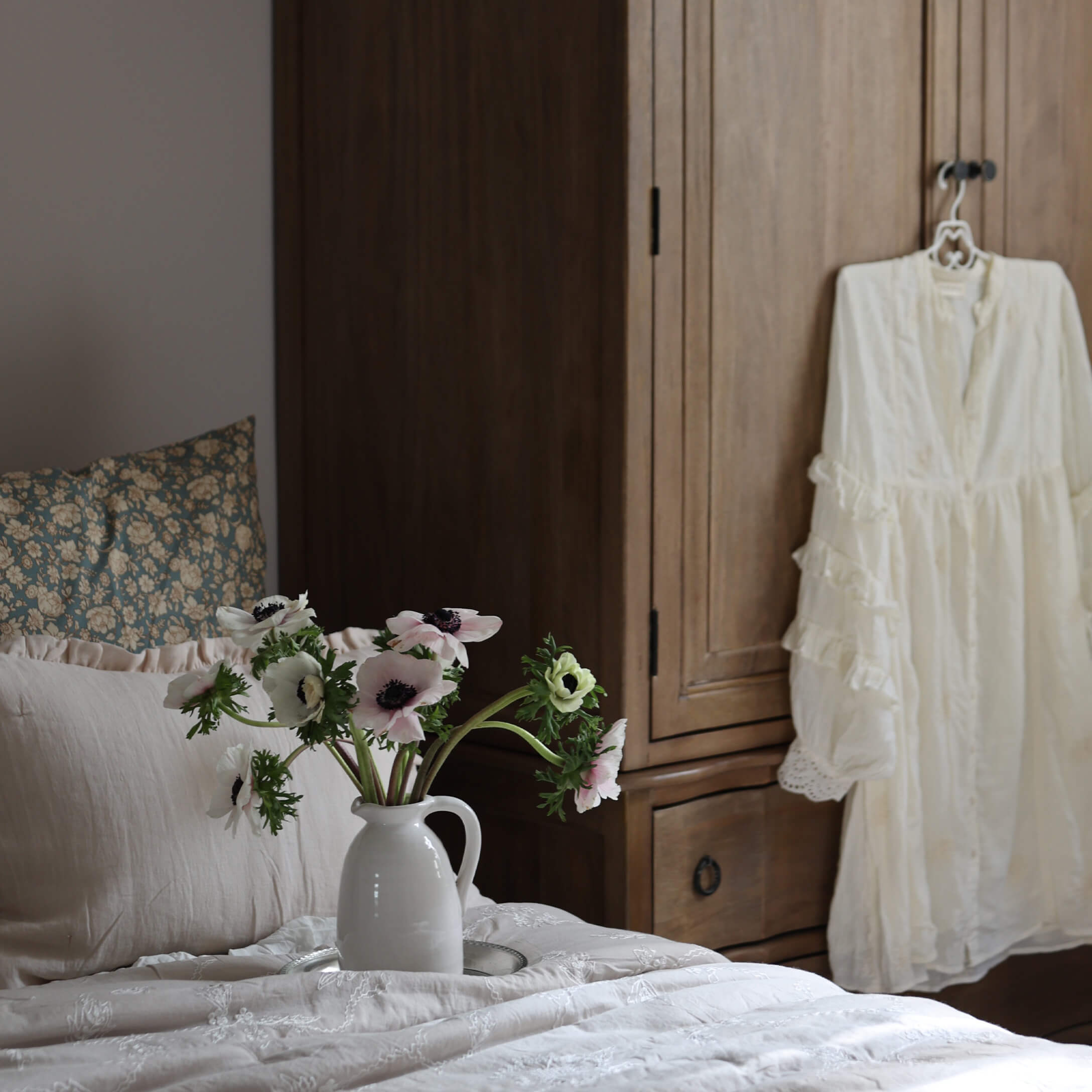 Weißer Keramikkrug mit Blumen auf hellem Bett vor Holzschrank im Schlafzimmer