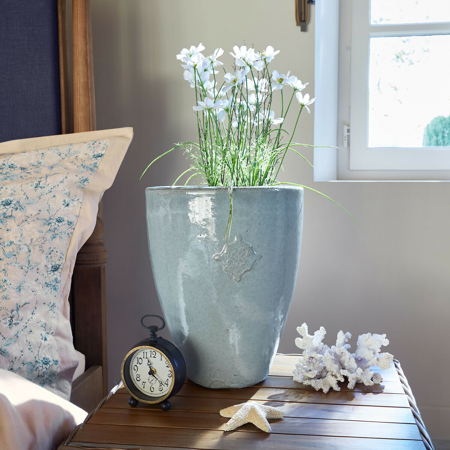 Grauer Keramikblumentopf mit weißen Blumen auf Holztisch neben Wecker und maritimen Dekorationen