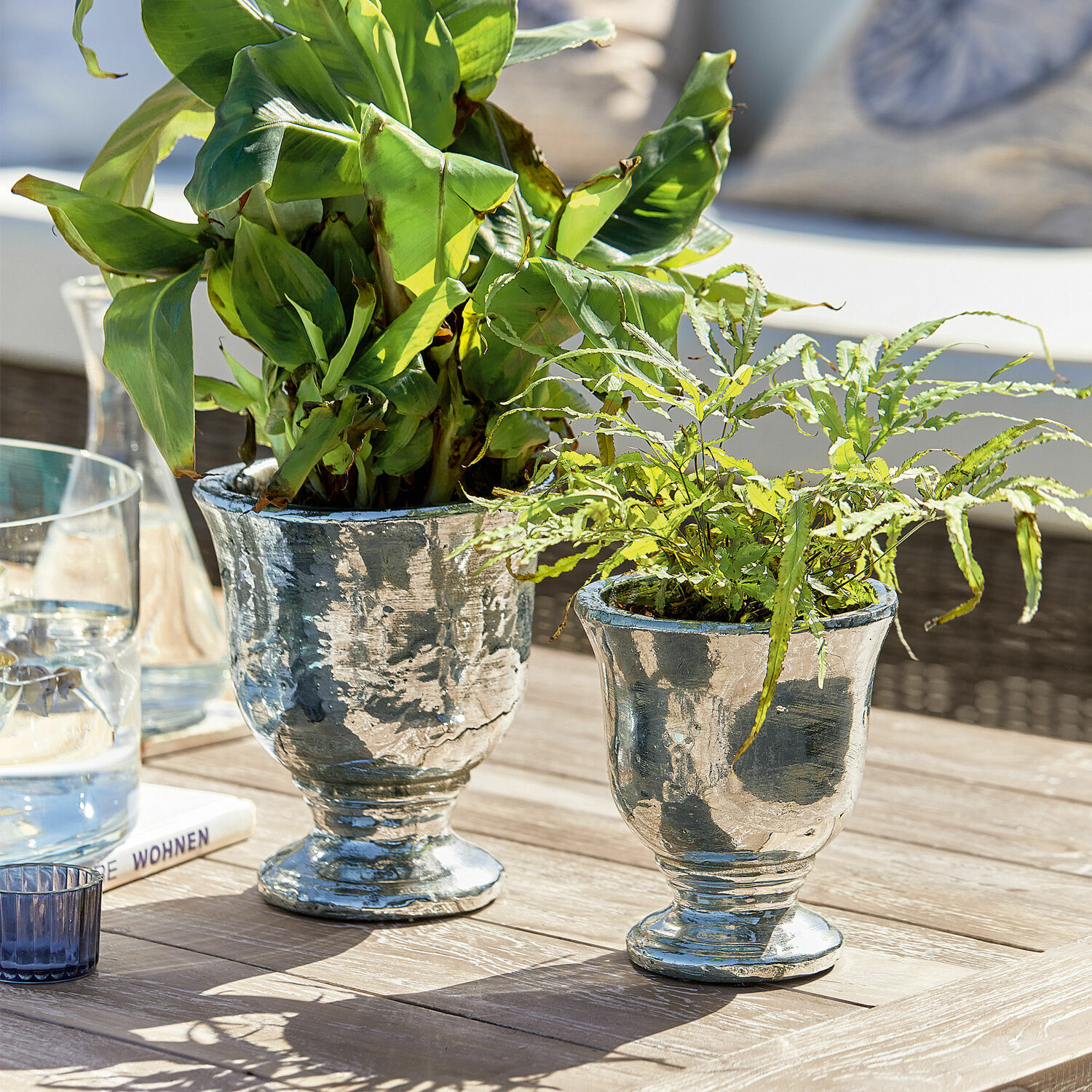 Zwei silberne Metall-Pflanzgefäße mit grünen Pflanzen auf Holztisch in hellem Ambiente