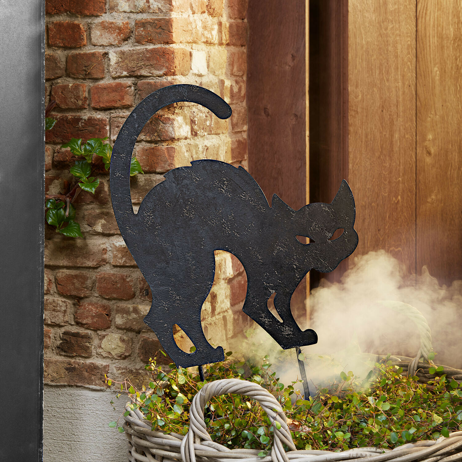 Gartenstab Rénelise Dekorative schwarze Metallkatzen-Silhouette in einem Korb mit grünen Pflanzen vor einer Ziegelwand