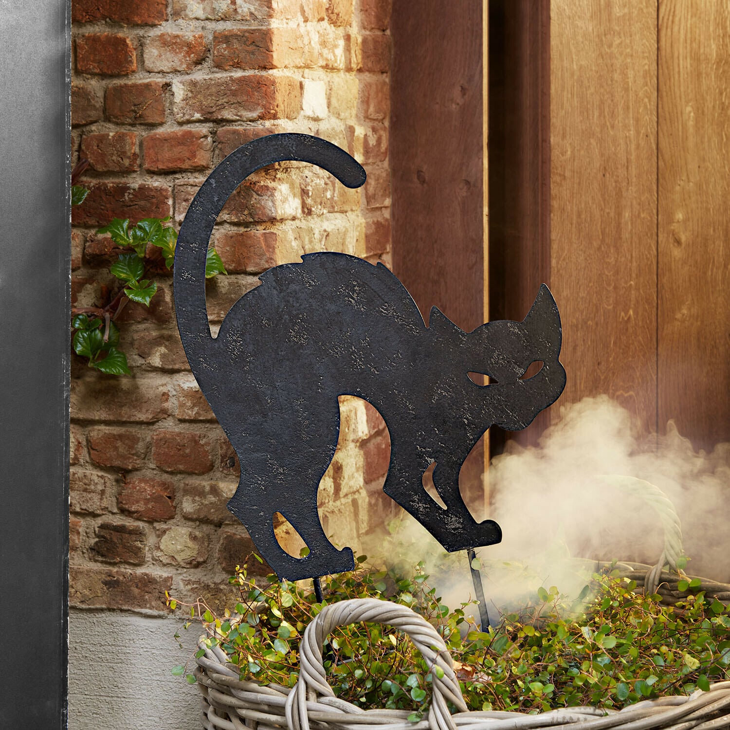 Gartenstab Rénelise Dekorative schwarze Metallkatzen-Silhouette in einem Korb mit grünen Pflanzen vor einer Ziegelwand