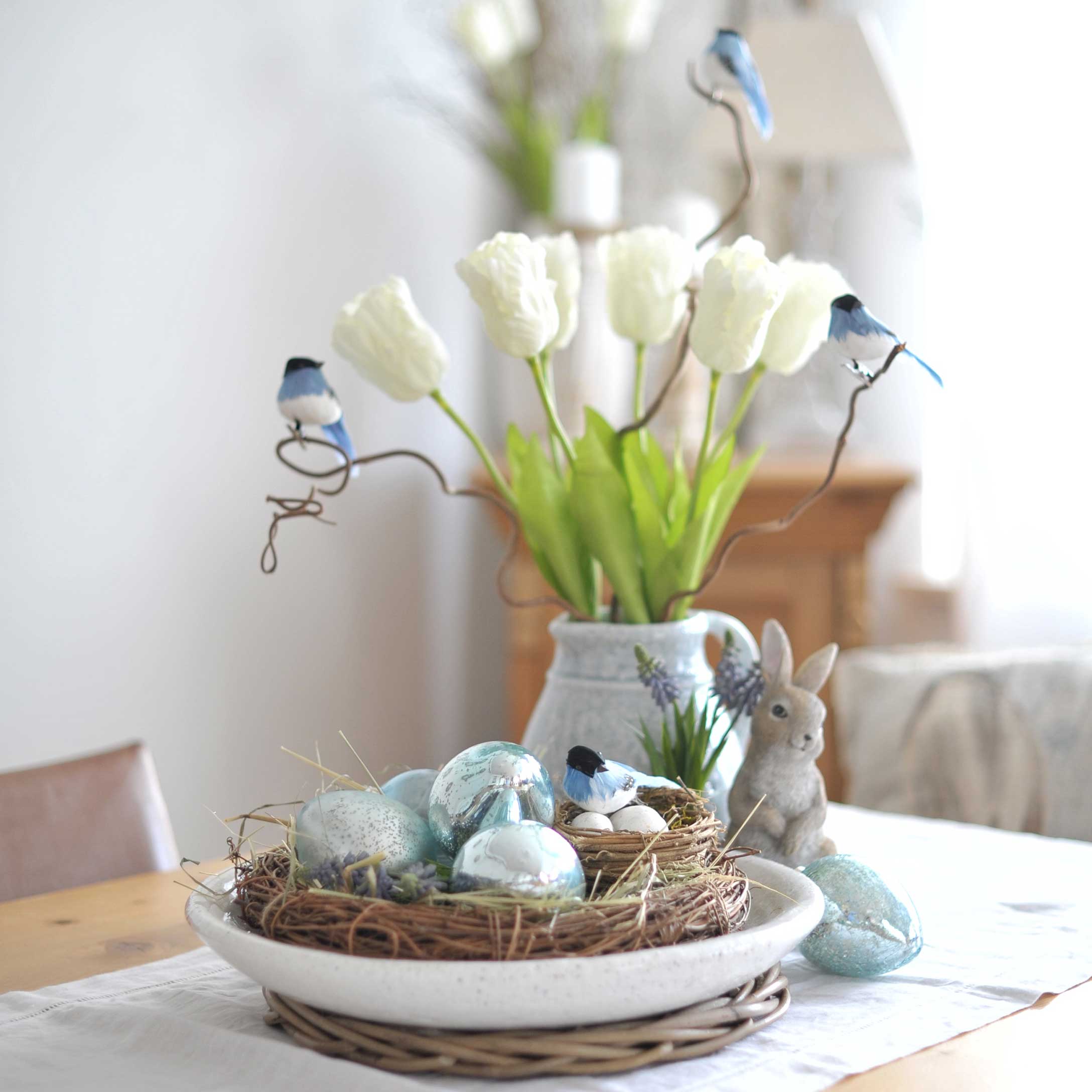 Dekorativer Kranz mit blauen Eiern und Vögeln auf Tisch mit weißen Tulpen und Hasenfigur