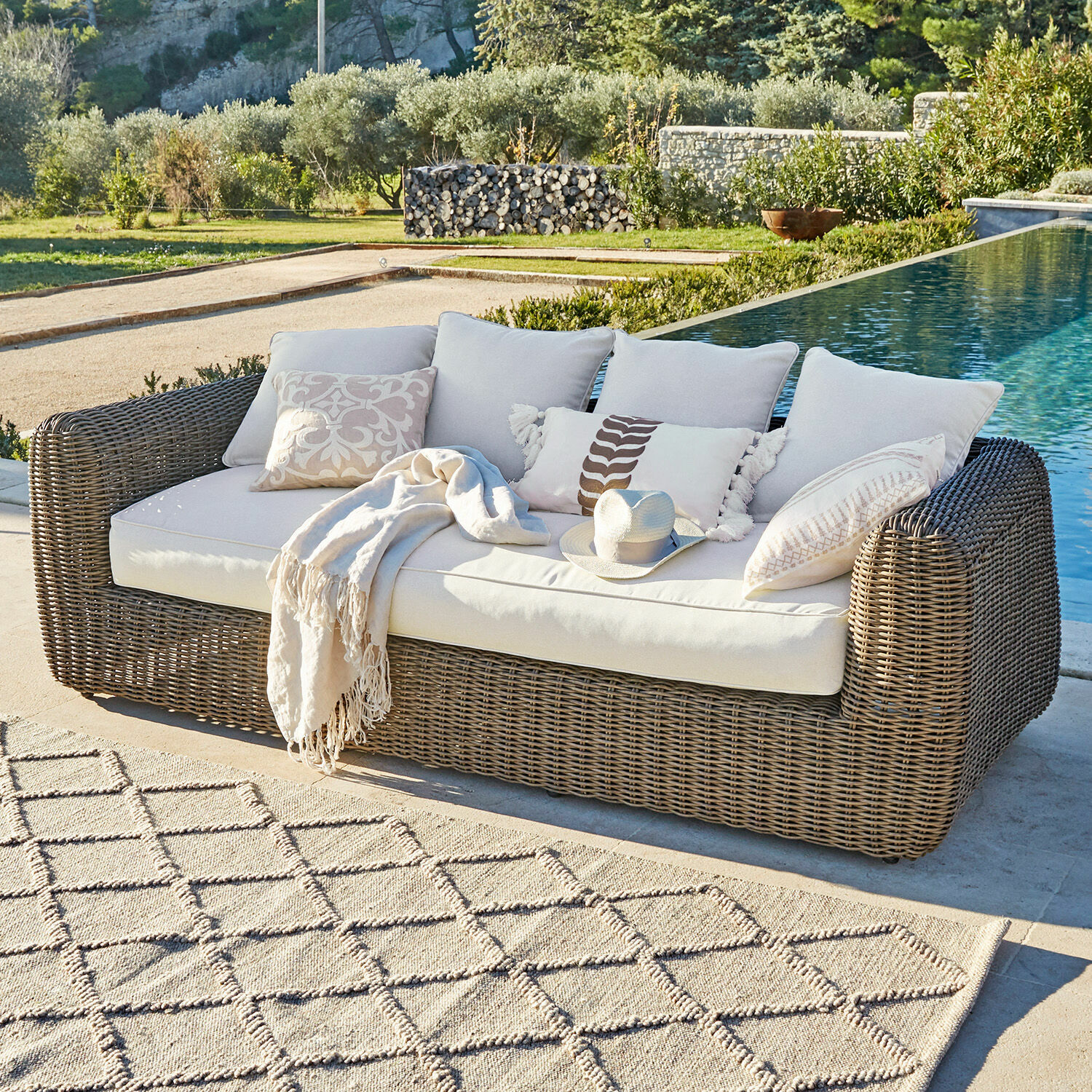 Gartensofa aus braunem Rattan mit cremefarbenen Kissen und Decke am Pool