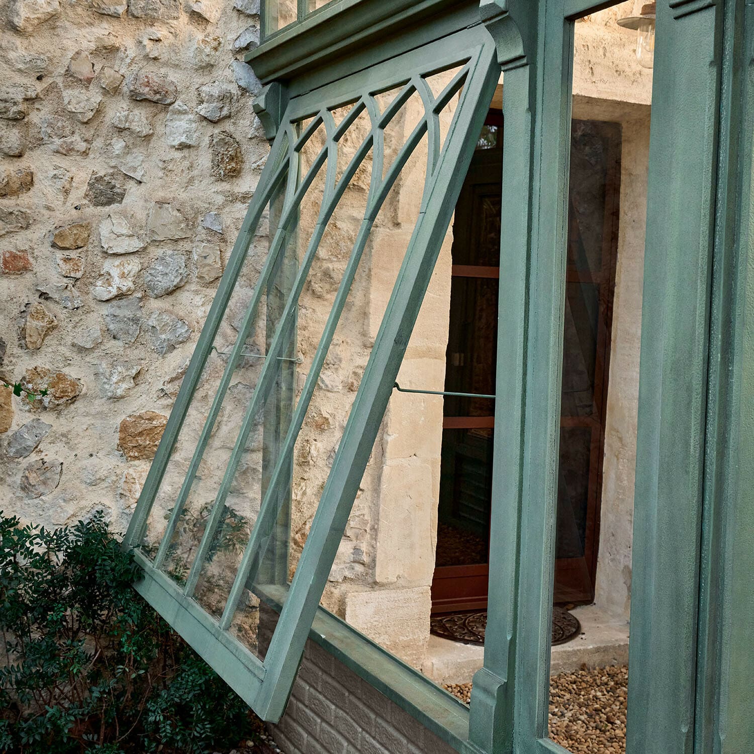 Grünes Metallfenster mit dekorativen Streben, nach außen geöffnet, vor Steinmauer und Pflanzen