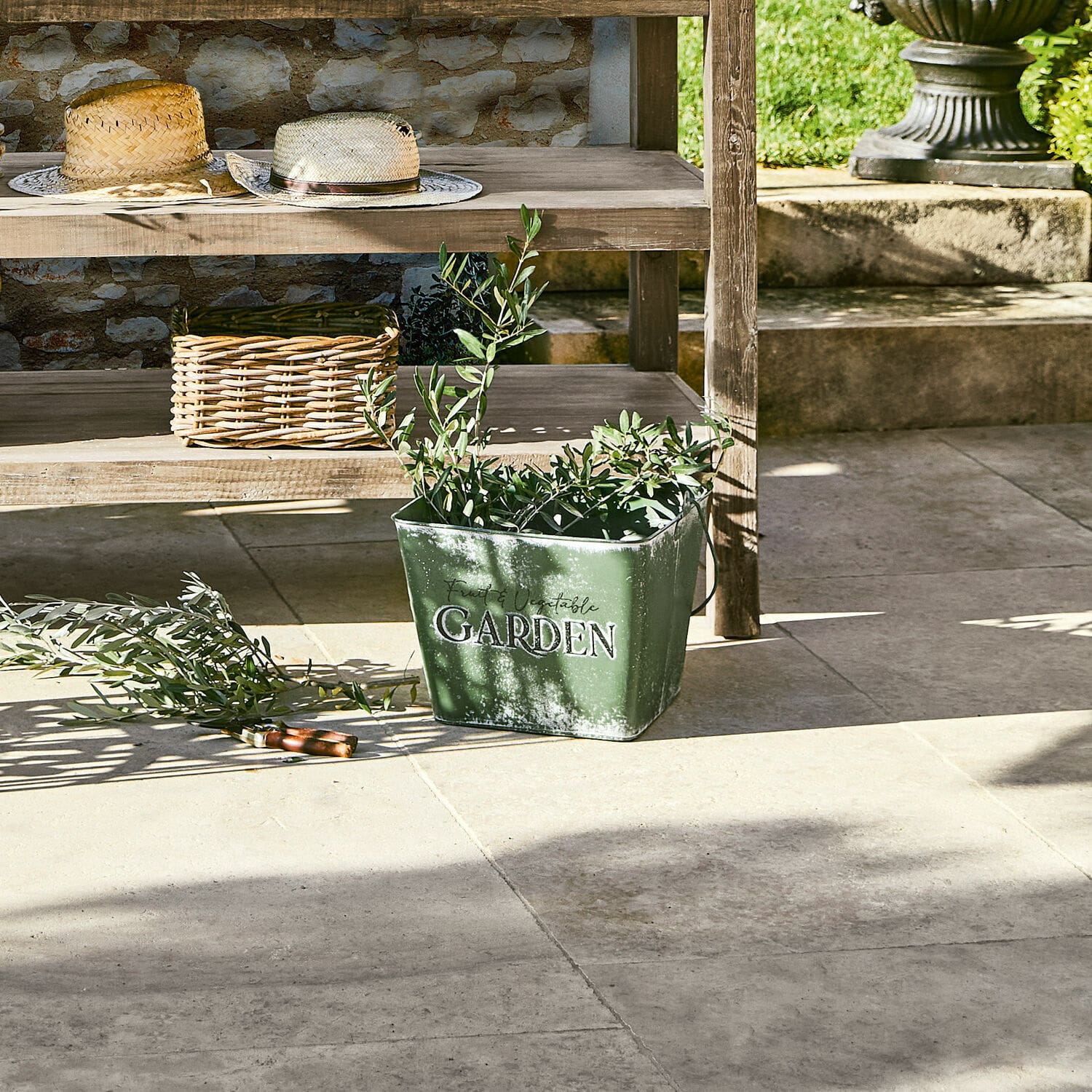 Grüner Metall-Eimer mit Aufschrift Garden und Pflanzen auf Steinboden im Garten