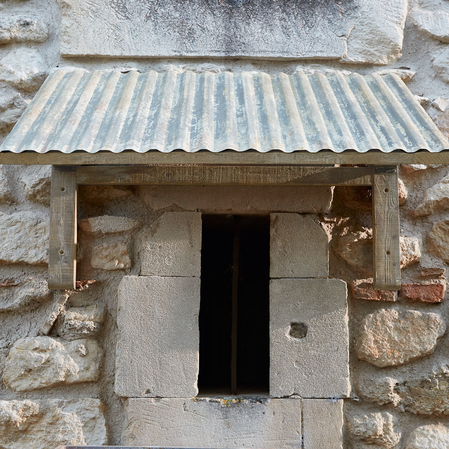 Kleines Wellblechdach mit Holzrahmen über einem steinernen Fenster in einer Steinmauer
