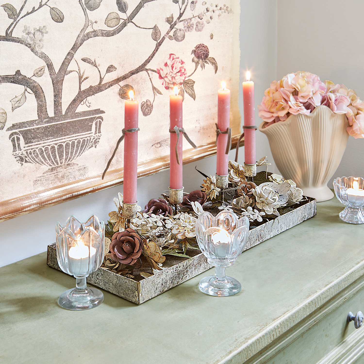 Dekorativer Kerzenhalter mit Metallblumen und rosa Kerzen auf grünem Sideboard mit Vase