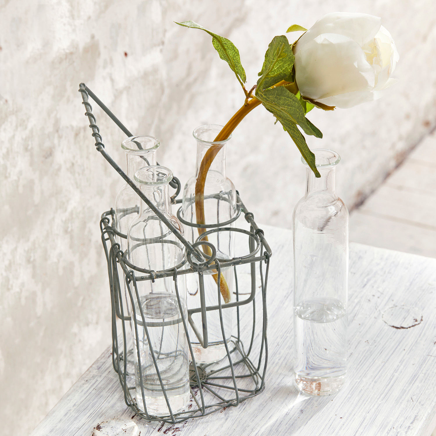 Glasflaschen mit Wasser im grauen Metallständer auf weißem Holztisch mit weißer Blume