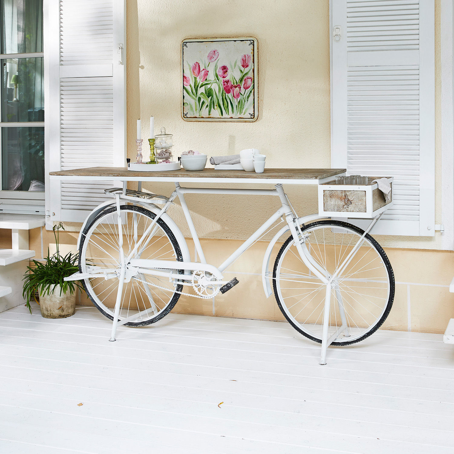 Weißer Metall-Fahrrad-Tisch mit Holzplatte, Dekorationen und Geschirr auf heller Terrasse