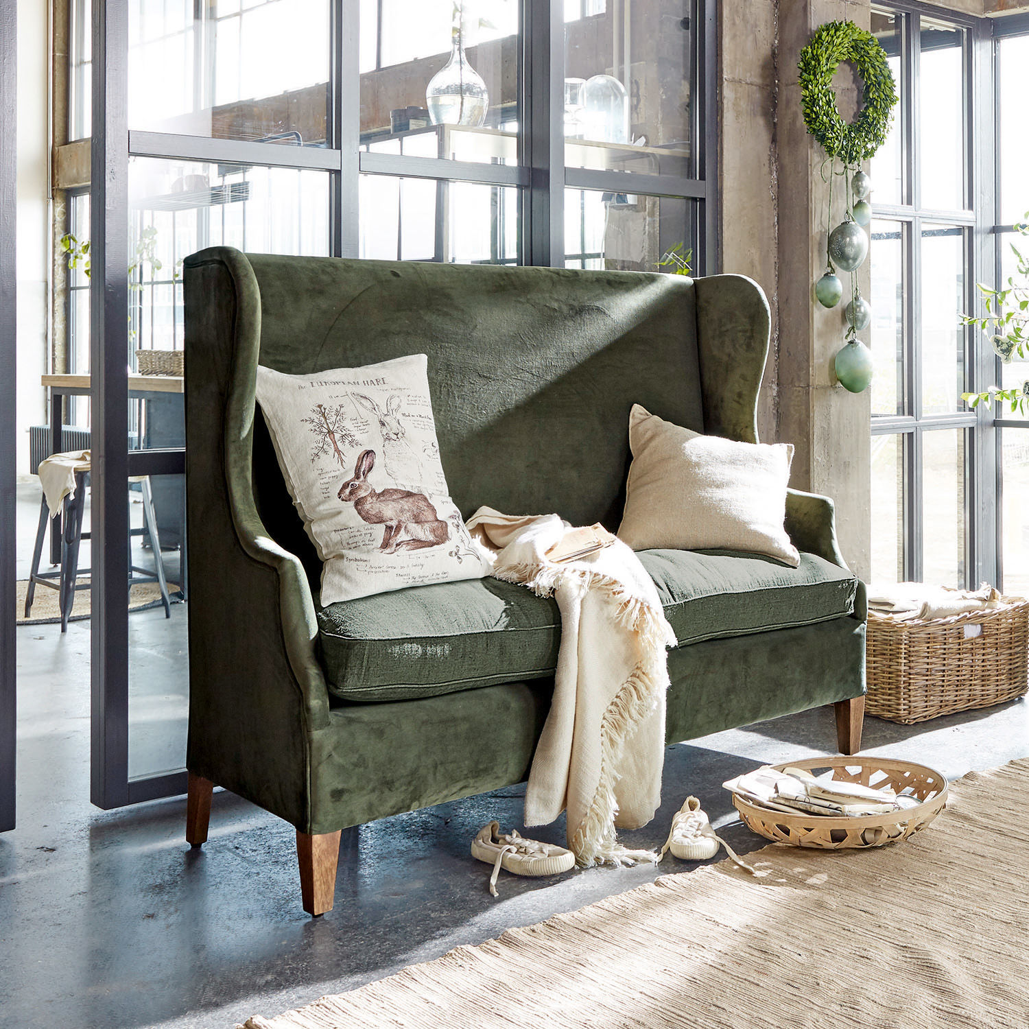 Sofa Ombra Grünes Samtsofa mit Kissen und cremefarbenem Plaid in hellem, modernem Raum mit großen Fenstern