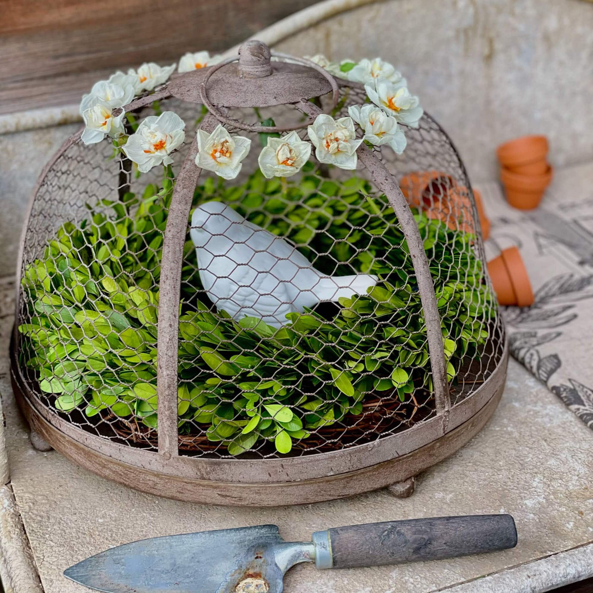Metall-Gartendekoration mit grünem Laub und weißem Keramikvogel unter blumengeschmücktem Käfig