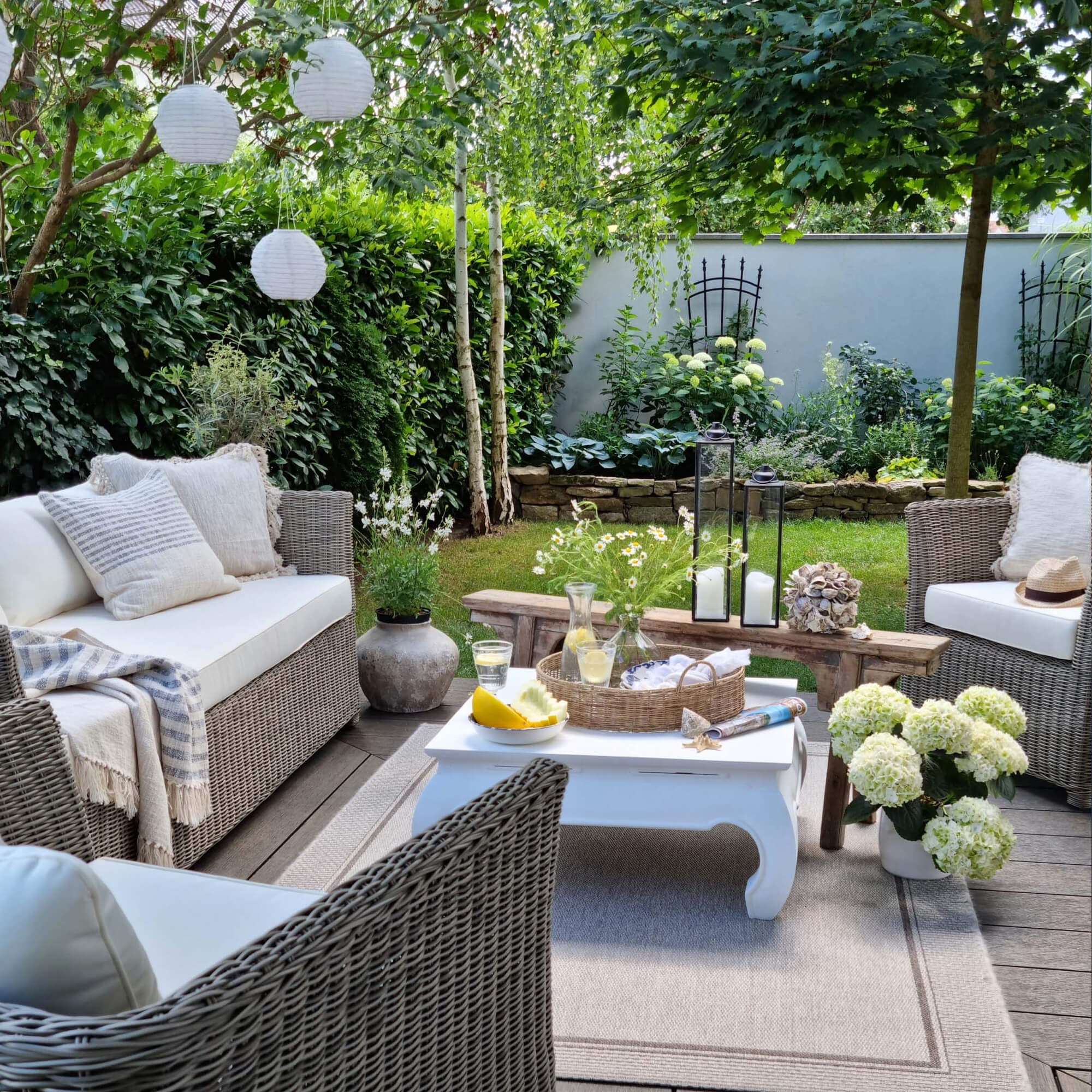 Gemütliche Gartenecke mit grauen Rattanmöbeln und weißen Kissen, Dekorationen und grünen Pflanzen.