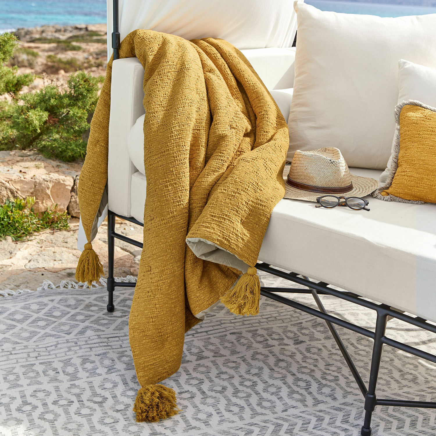 Plaid Rabea Gelbe Tagesdecke mit Quasten auf weißem Outdoor-Sofa, Terrasse mit Sonnenbrille und Hut dekoriert
