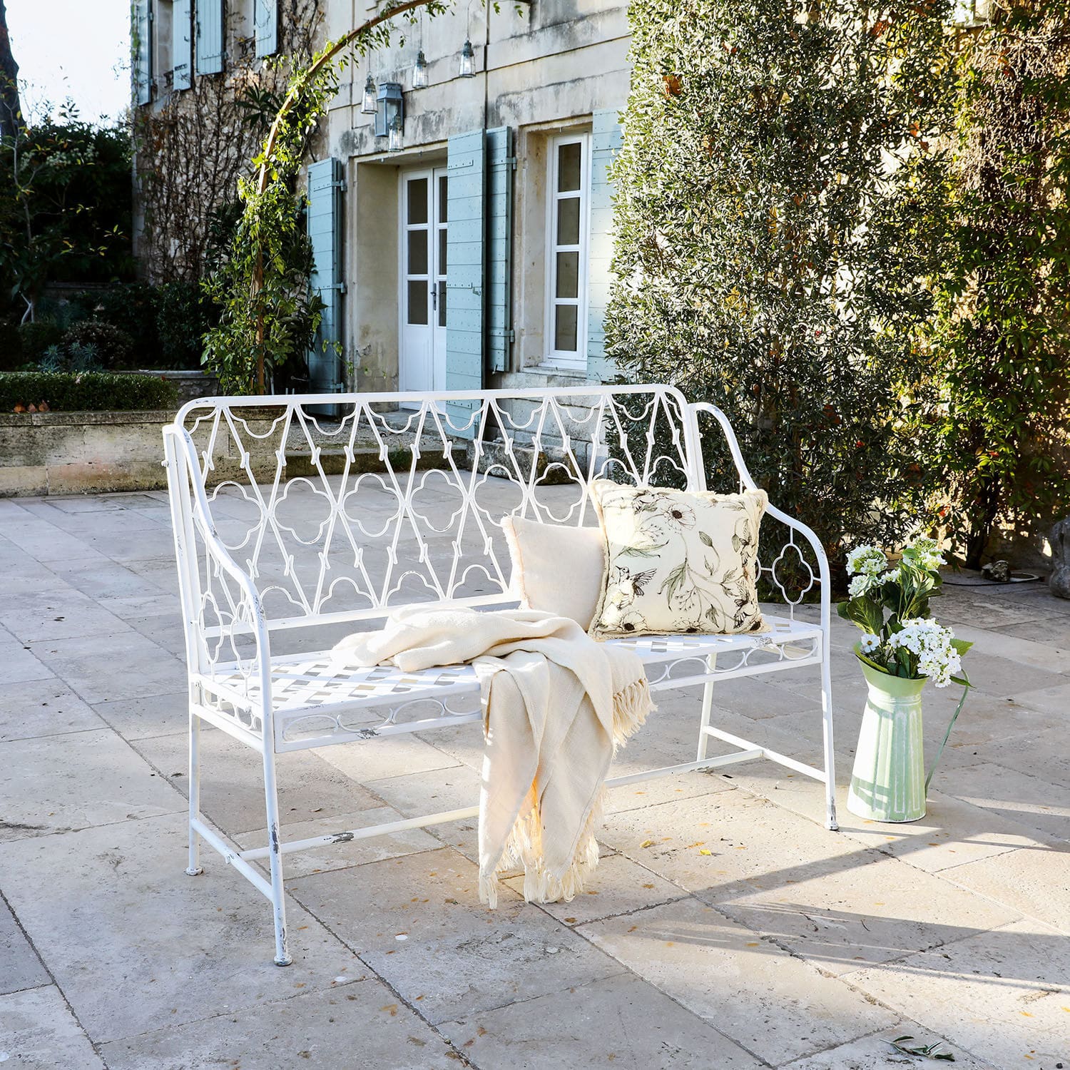 Weiße Metall-Gartenbank mit Kissen und Decke auf Stein-Terrasse neben grünen Pflanzen