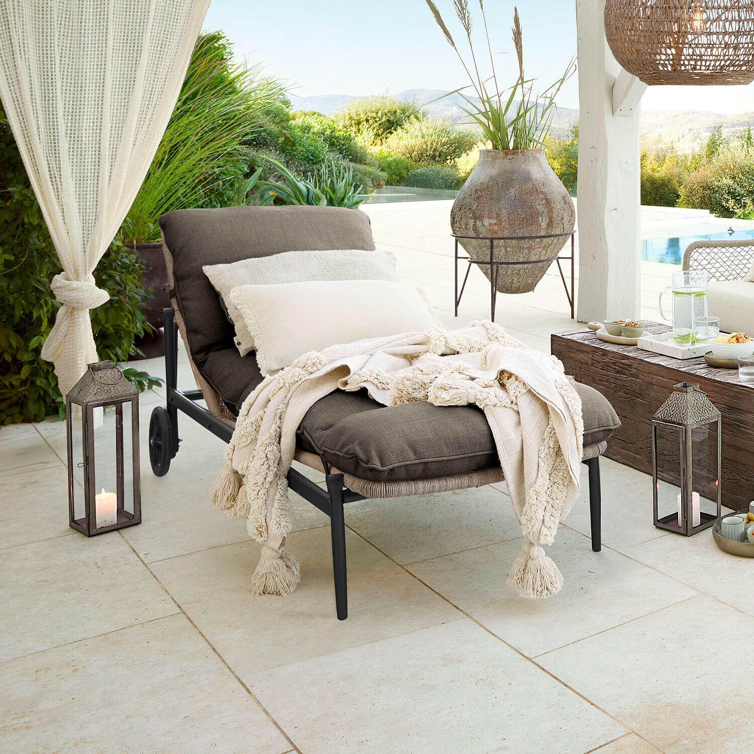 Gartenliege mit dunkelgrauem Stoff und cremefarbenem Fransenplaid auf Terrasse mit Gartenblick