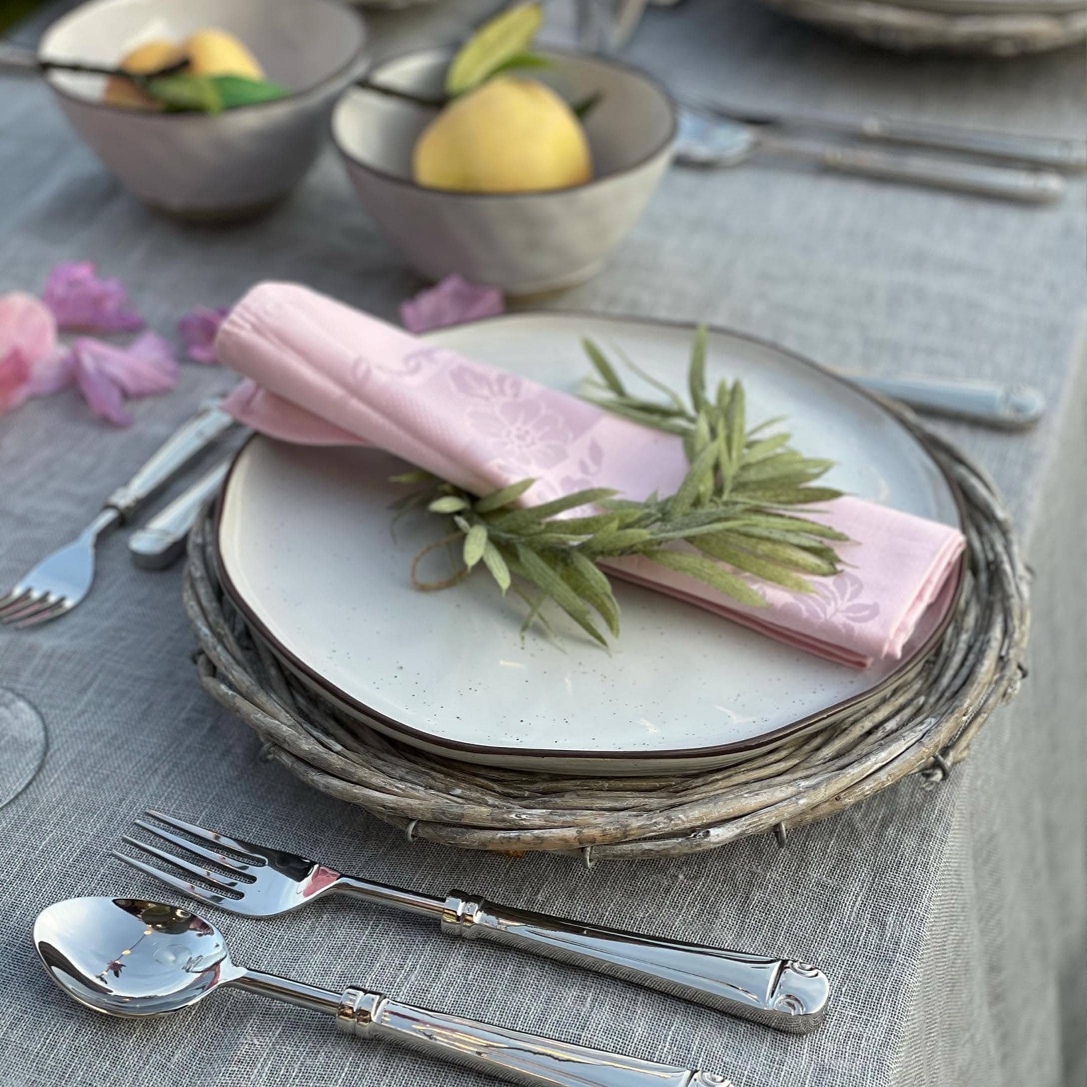 Elegantes Gedeck mit weißem Teller auf Weidenuntersetzer und rosa Serviette mit grünen Blättern