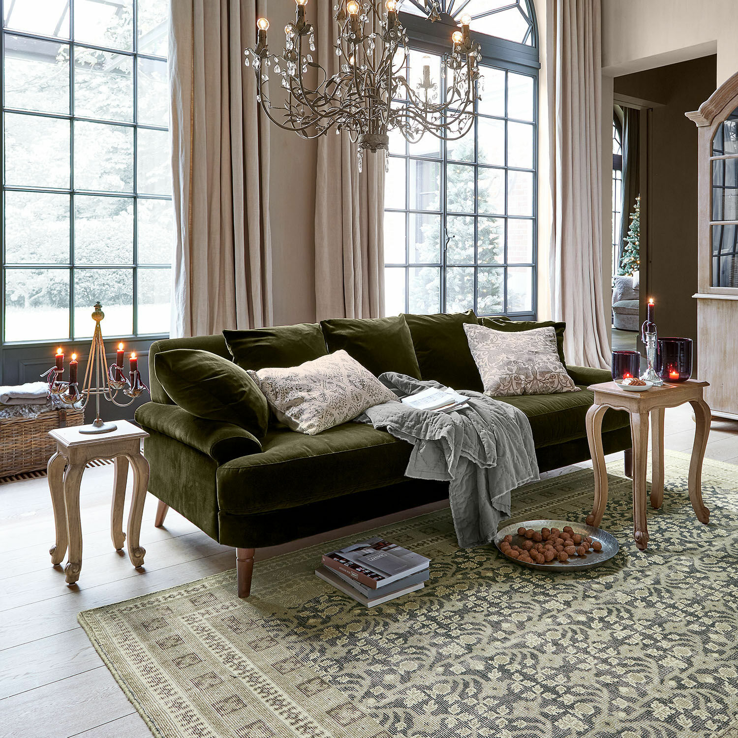 Sofa Rivermere Grünes Samtsofa mit Kissen und grauer Decke in hellem Wohnzimmer mit großen Fenstern